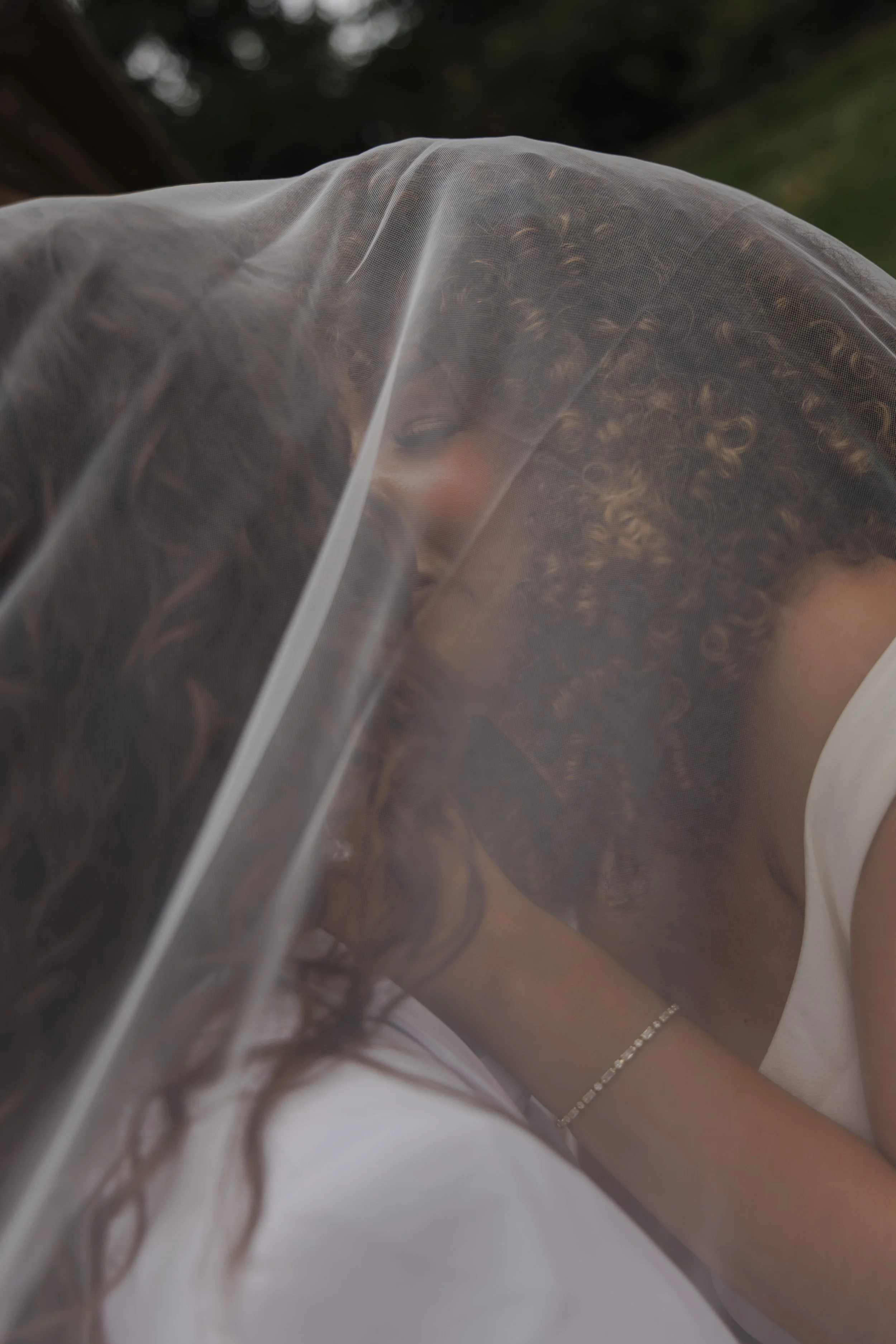 Close-up of a person with curly hair wearing a white top and a delicate bracelet on their arm, partially covered by a transparent plastic sheet, with blurred outdoor background.