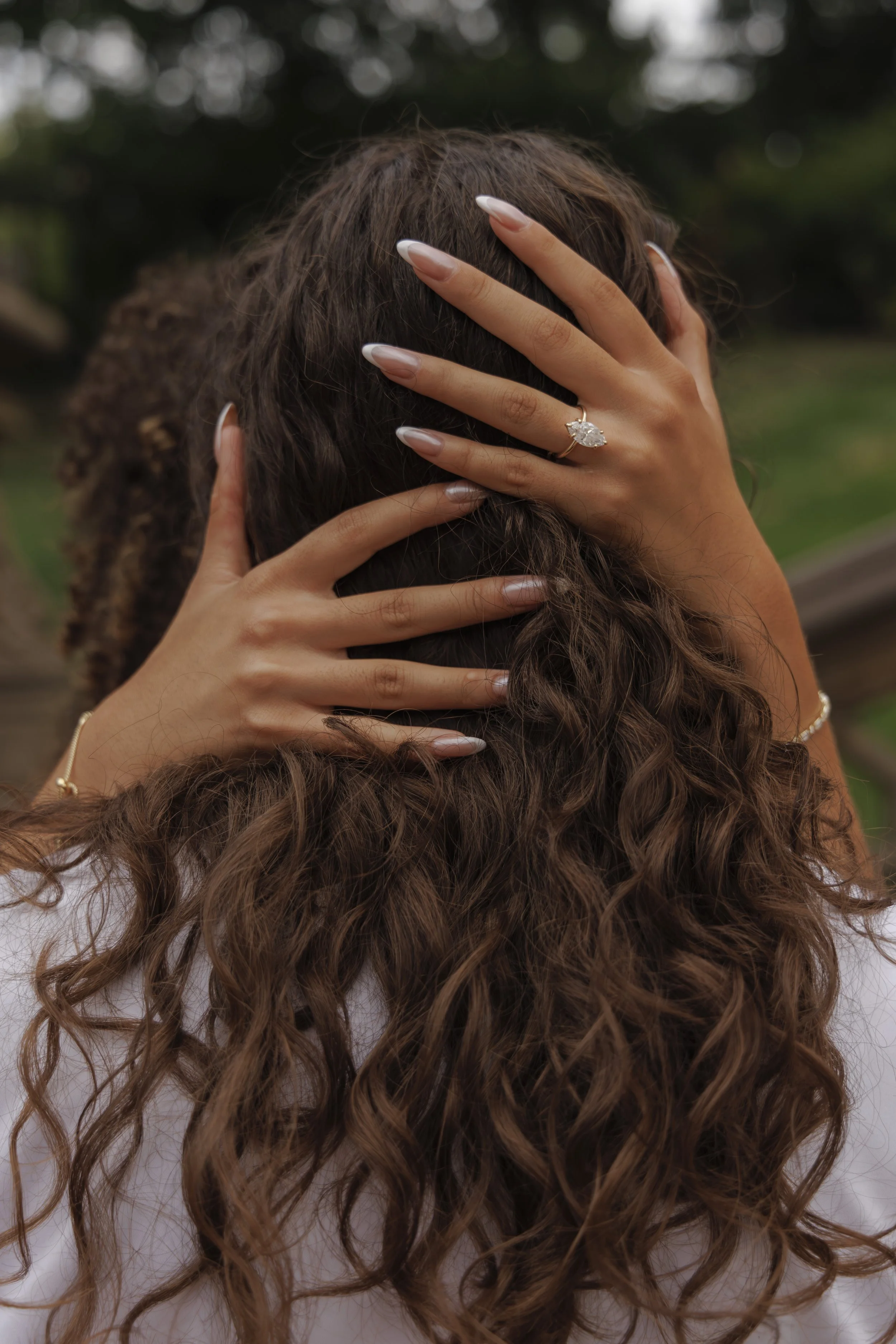 Close-up of a woman's head with long, curly brown hair, hands placed on her head, and a large diamond ring on her finger, outdoors with blurred greenery in the background.