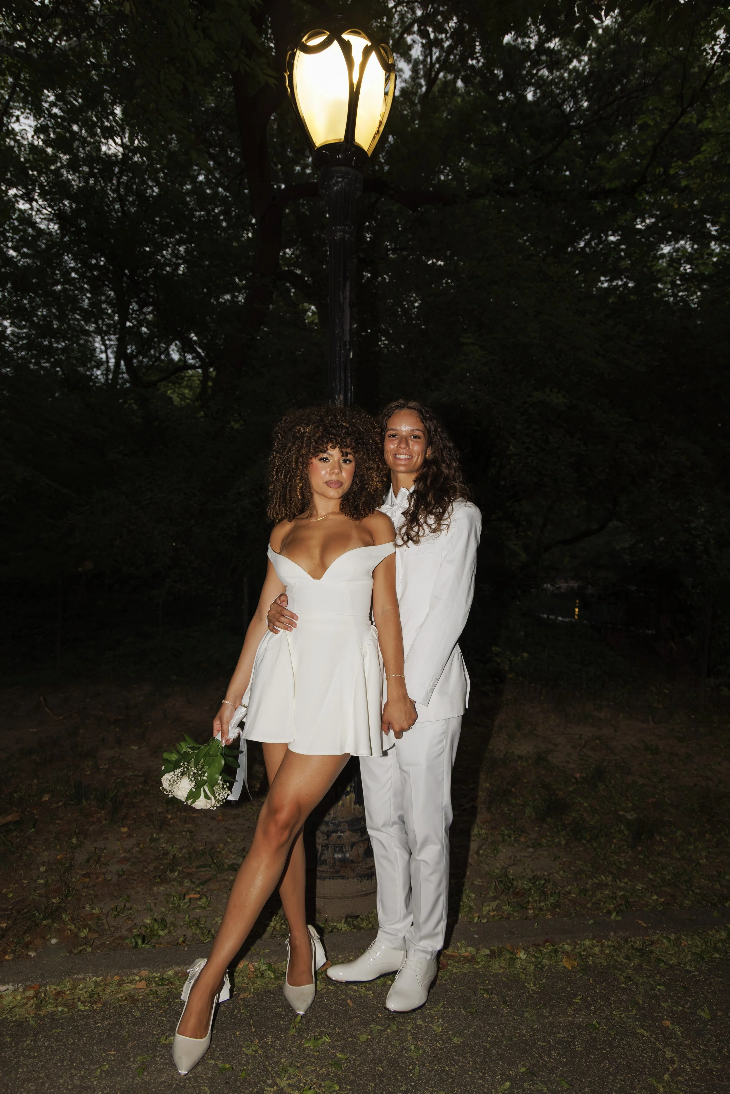 Two women standing close together outdoors at night, posing under a streetlamp. One woman is wearing an off-shoulder white dress and holding a small bouquet, the other woman is dressed in a white suit with a white shirt. Both are smiling.