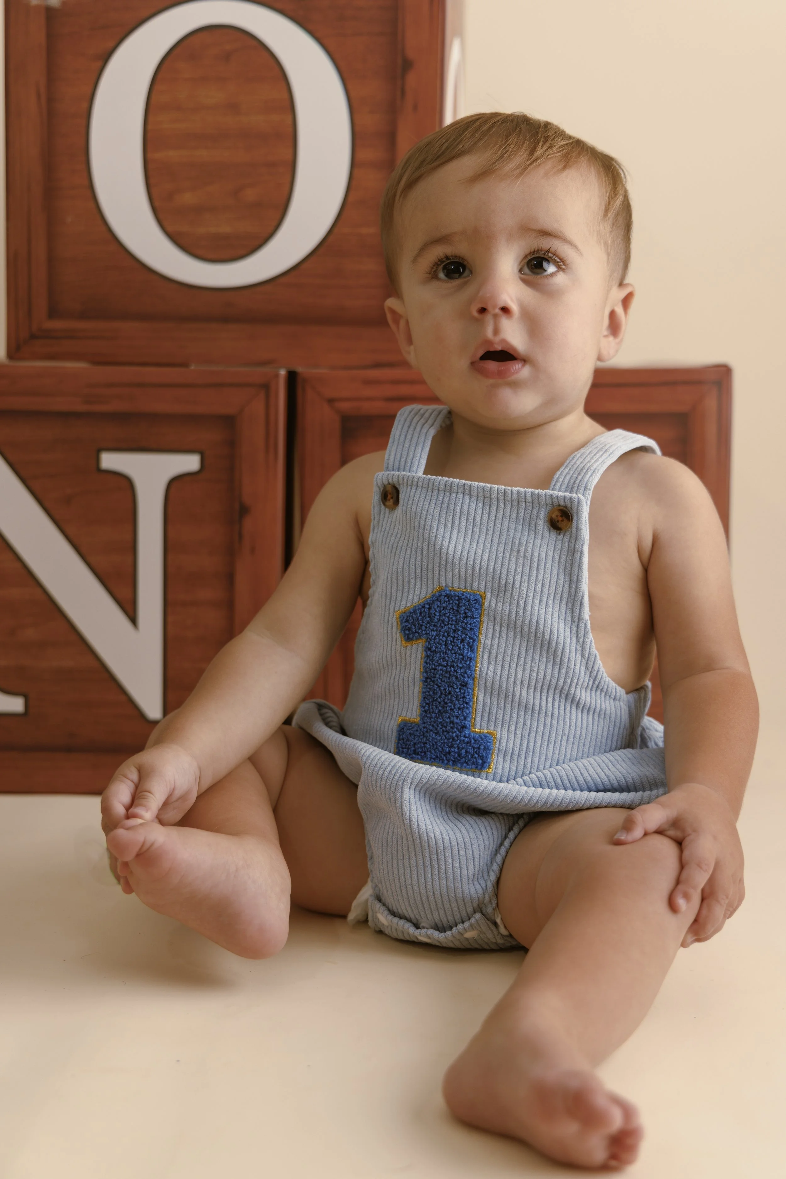 Young boy sitting on the floor in front of wooden blocks with the letter 'O' and 'N' visible, wearing a sleeveless gray onesie with a blue number one on it and looking up with a surprised expression.