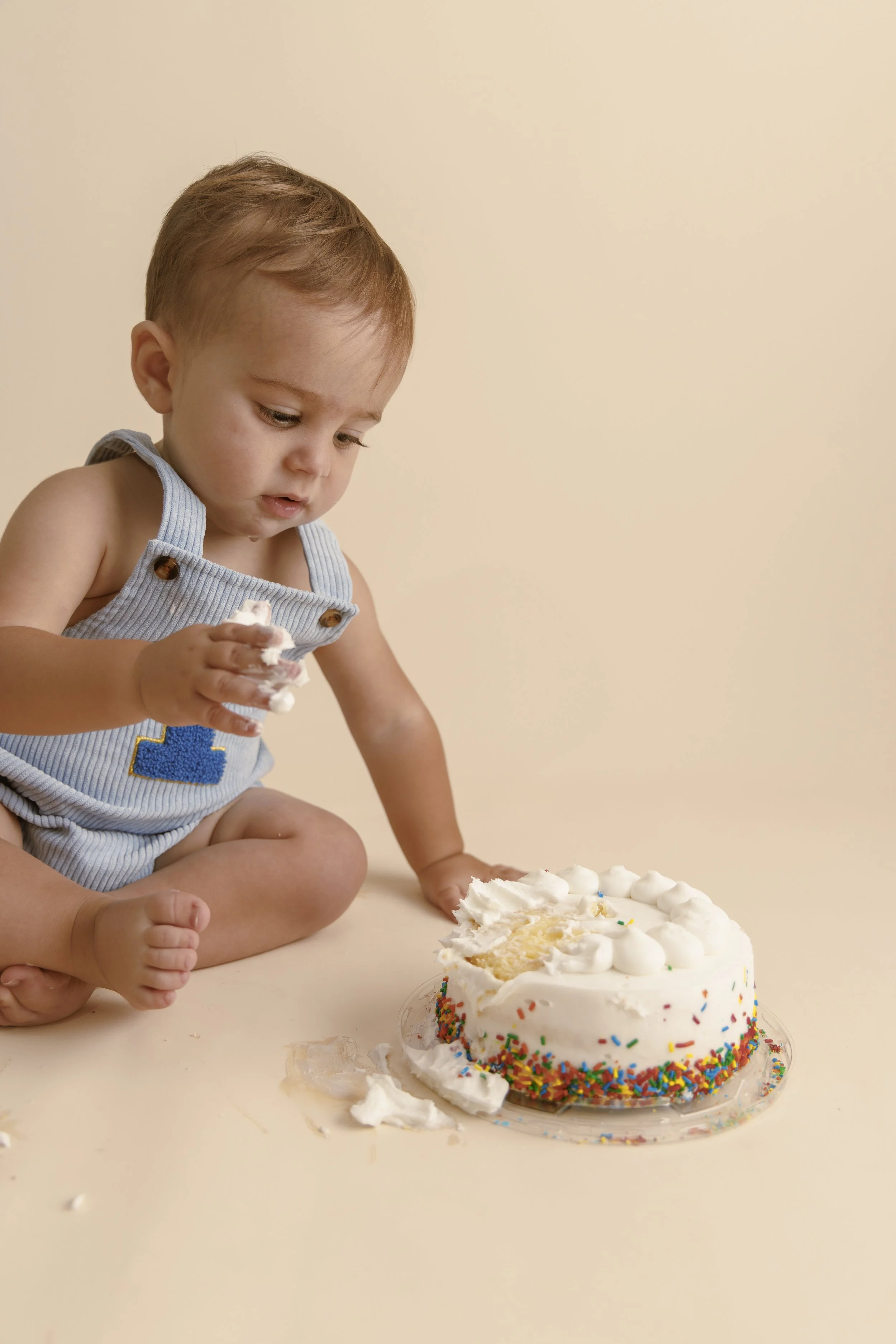 A young child with light brown hair, wearing blue overalls, is sitting on the floor and holding a piece of cake. There is a birthday cake with white frosting, colorful sprinkles, and some cake missing in front of the child. The background is plain and light-colored.