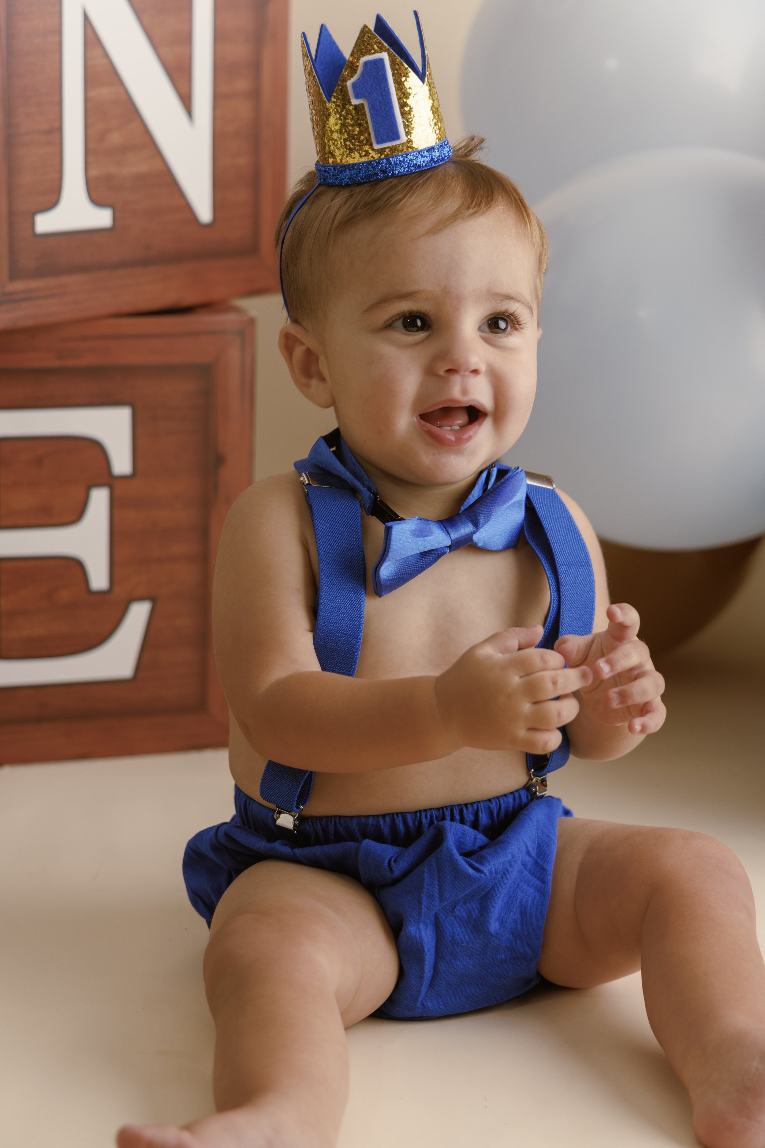 A young boy wearing blue suspenders, shorts, and a blue bow tie, sitting on the floor with a birthday crown that has a number 1 on it. There are large blocks with letters in the background and a white balloon to his right.