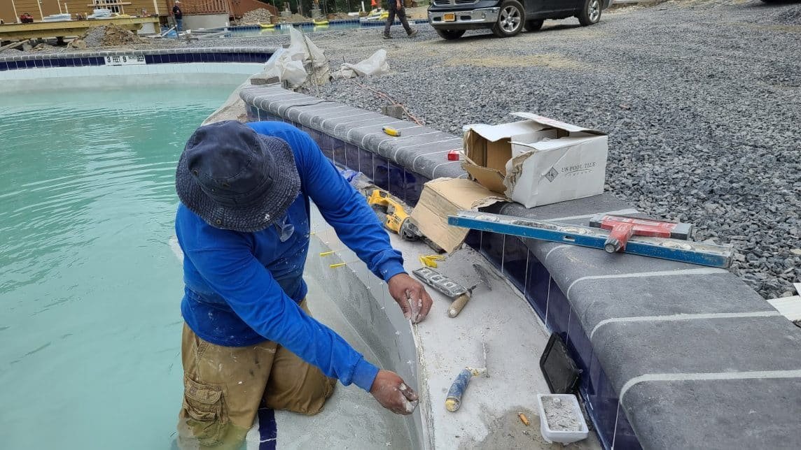 A worker in blue shirt and khaki pants kneels by a pool, installing or repairing the pool's edge with tools and materials nearby.