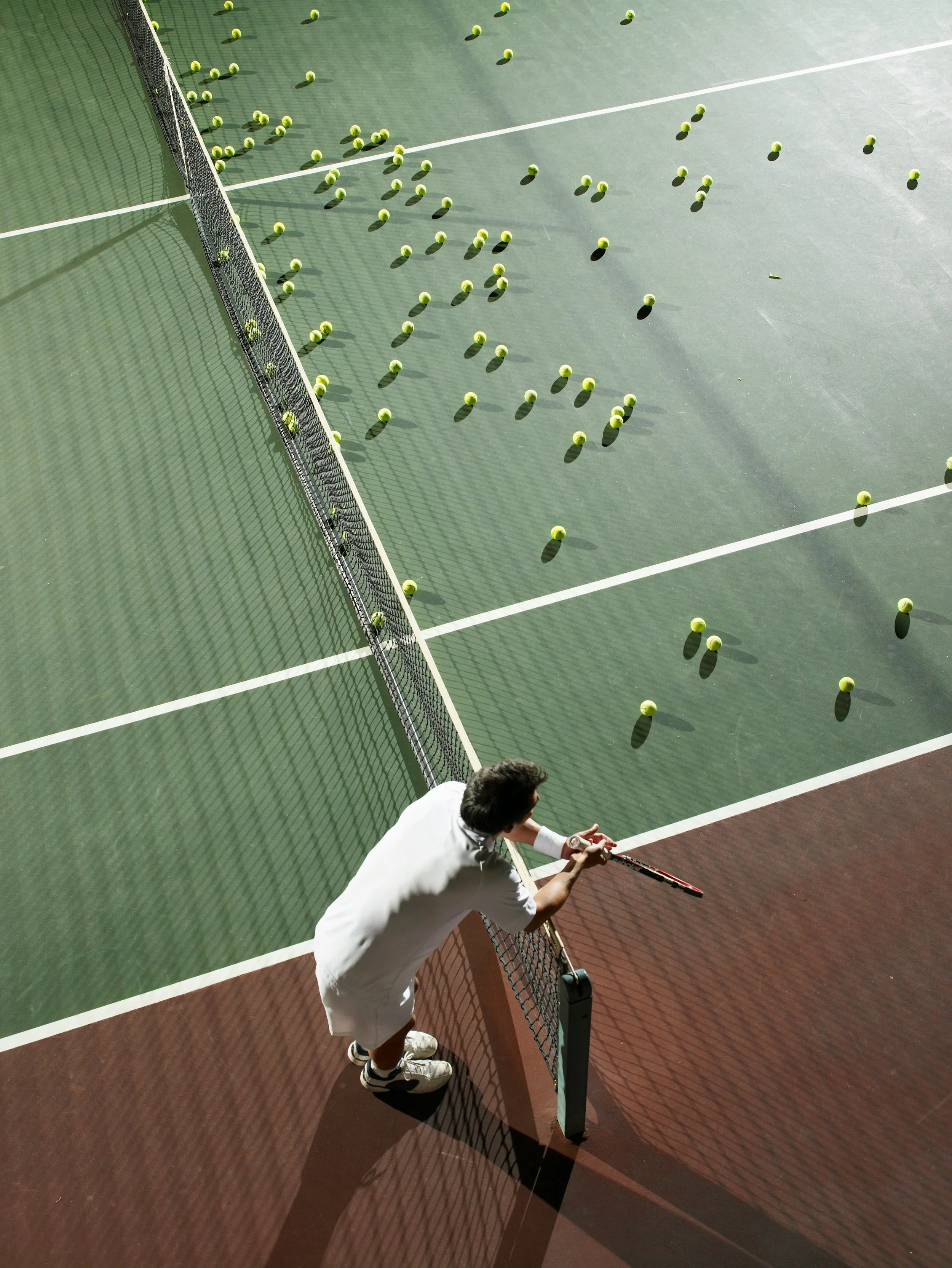 Man leaning on a tennis net on a tennis court, looking at scattered tennis balls on the court surface.  Feeling gassed after a long training session is easily avoidable with Game Day Fuel.  Clean ingredients boost stamina and mental focus.