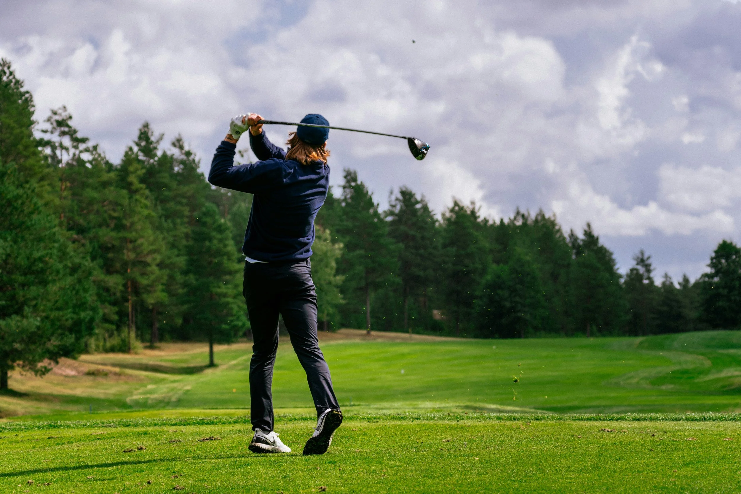 Man is swinging a golf club on a golf course with green grass, trees, and a cloudy sky in the background.  Playing a round of golf requires extreme focus.  Game Day Fuel helps with the proper nutrition.