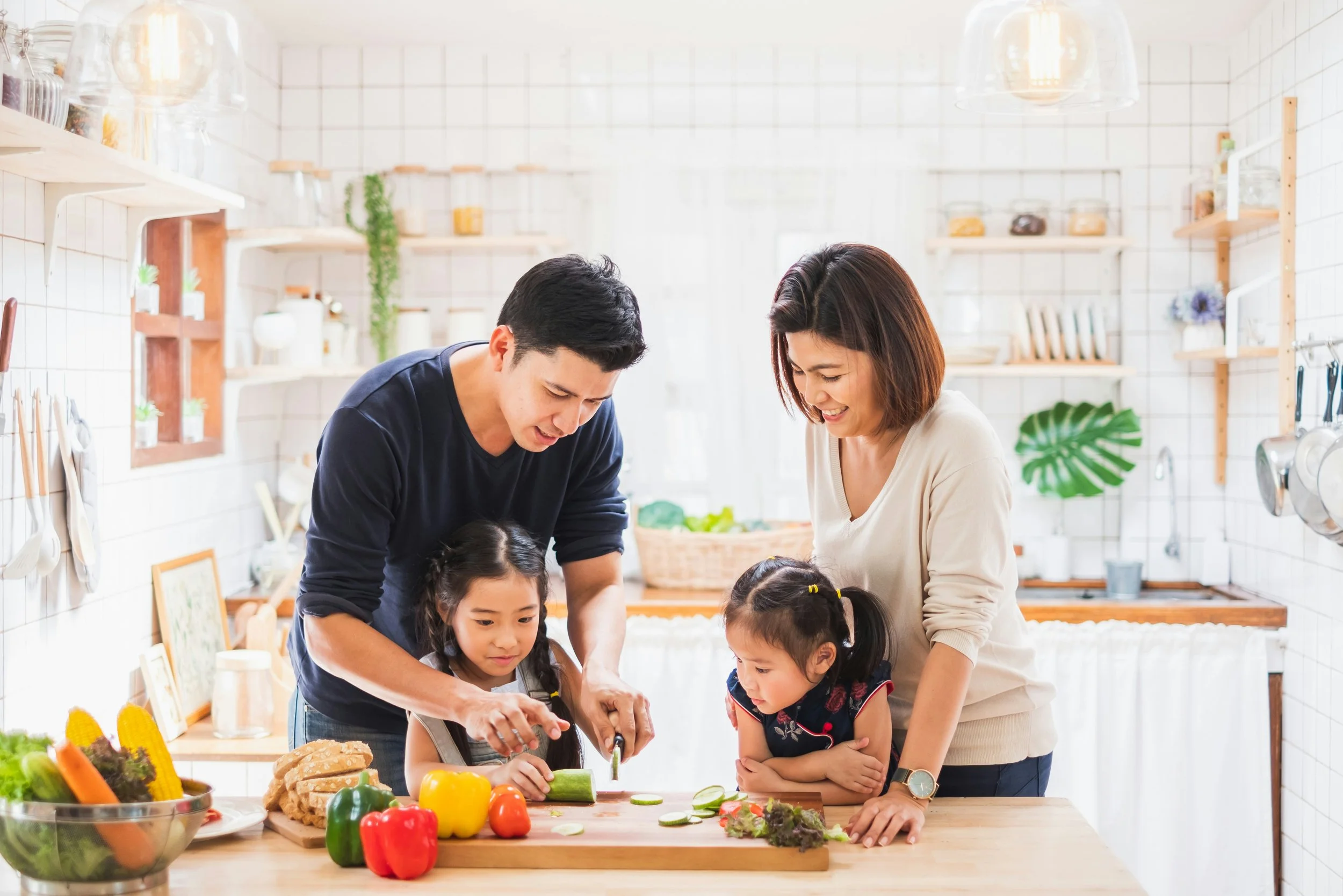 A family of four, including two children, cooking together in a bright kitchen with white tile walls, preparing vegetables on a wooden countertop.