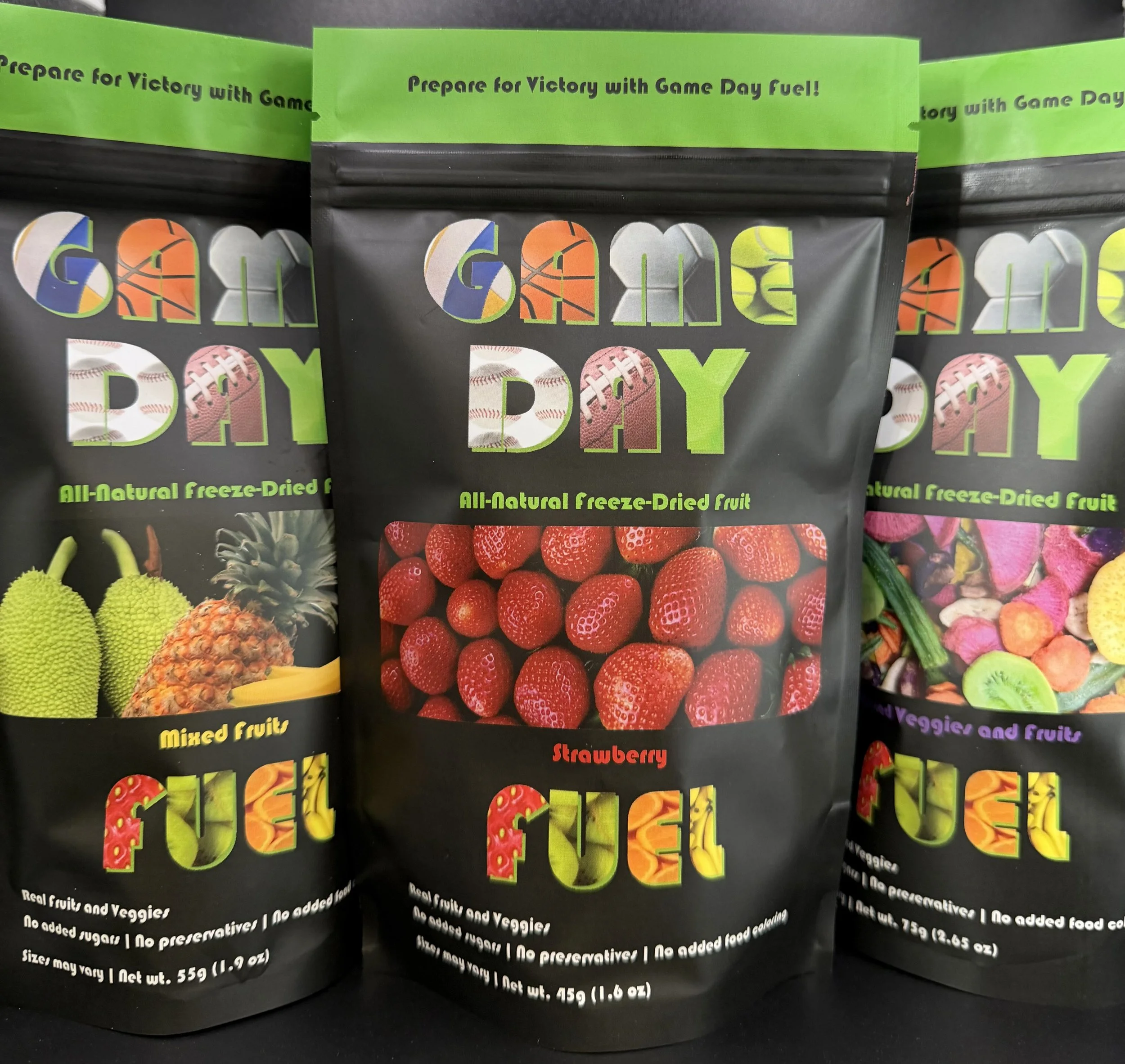 Black packaging of freeze-dried fruit snacks labeled 'Gamer Day' with colorful letters, featuring images of strawberries in the center and assorted fruits on the side, with green header text 'Prepare for Victory with Game Day Fuel!' and descriptions of the product.