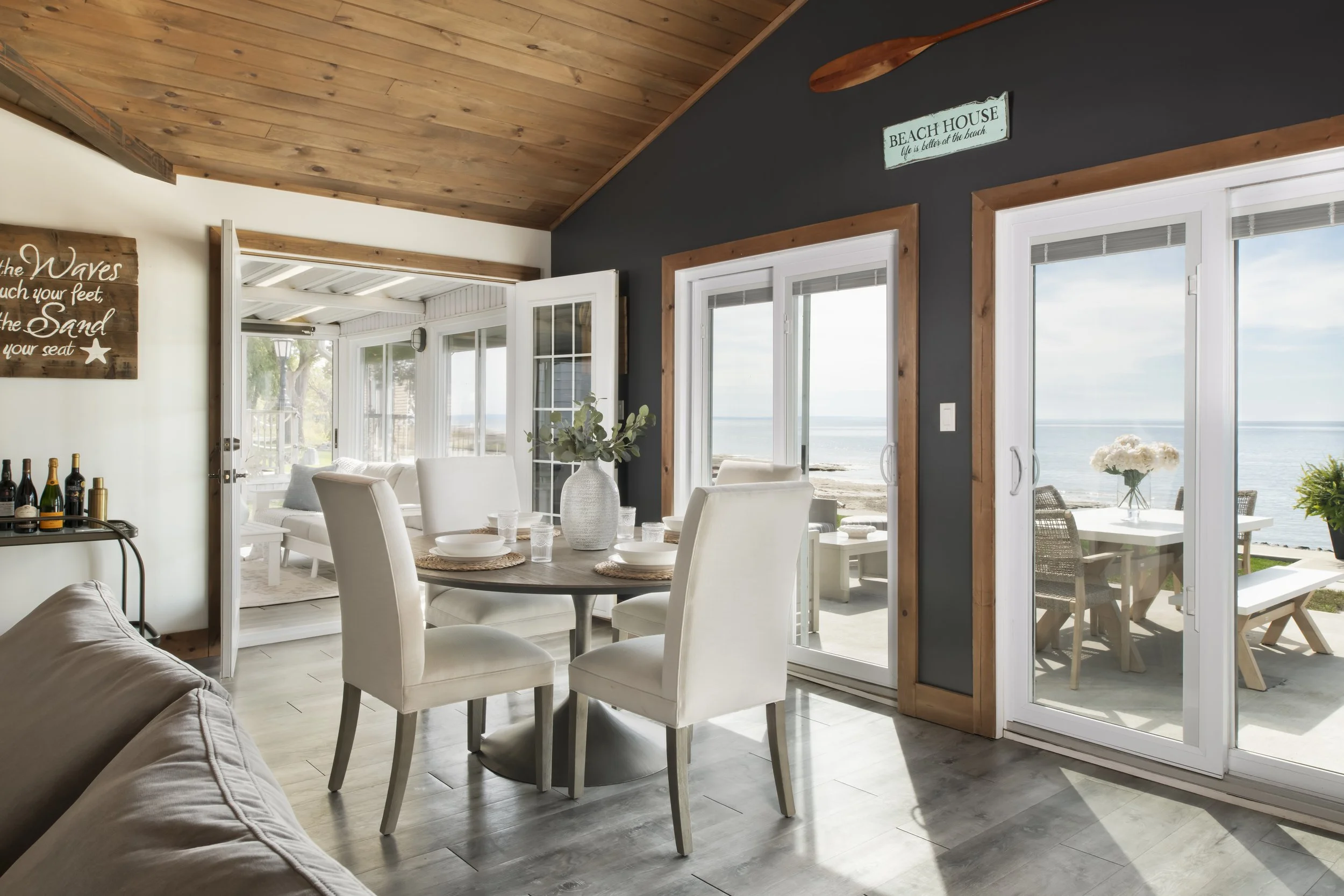 Beach house dining room with sliding glass doors leading to an outdoor patio and beach view, decorated with coastal signs, a round dining table with white chairs, and a vase of greenery.