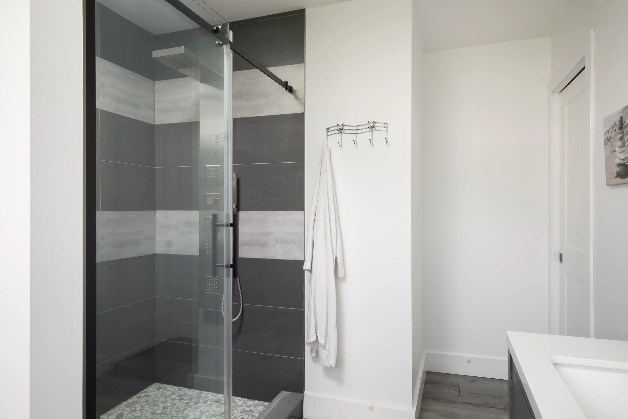Modern bathroom with a glass shower enclosure, dark gray tiles, a white wall with hooks holding a white towel, and a part of a white vanity on the right.