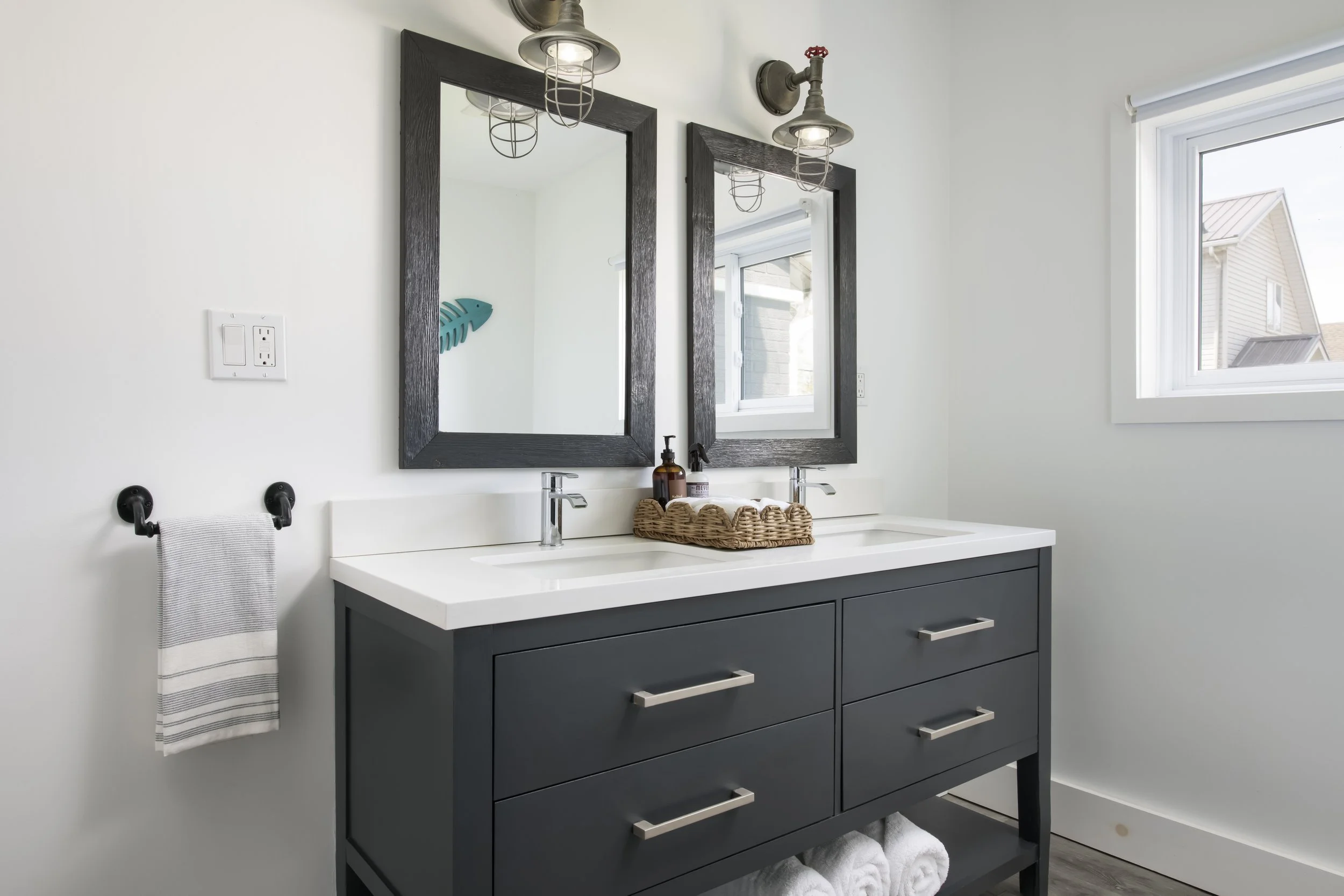 Modern bathroom vanity with dark gray cabinet, white countertop, two sinks, and black handles. Two mirrors with black frames above. Two industrial-style wall sconces. Towels in a basket and bathroom items on the counter. A window on the right side sh