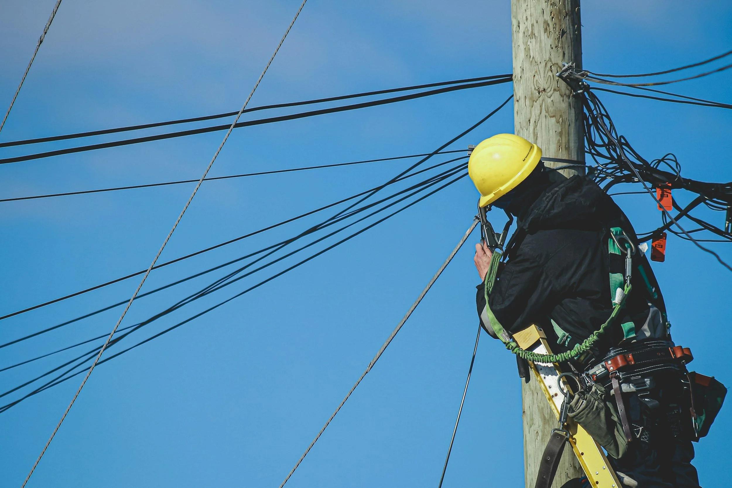 A worker in safety gear, including a yellow helmet and harness, is climbing or working on a utility pole with multiple power lines against a blue sky.