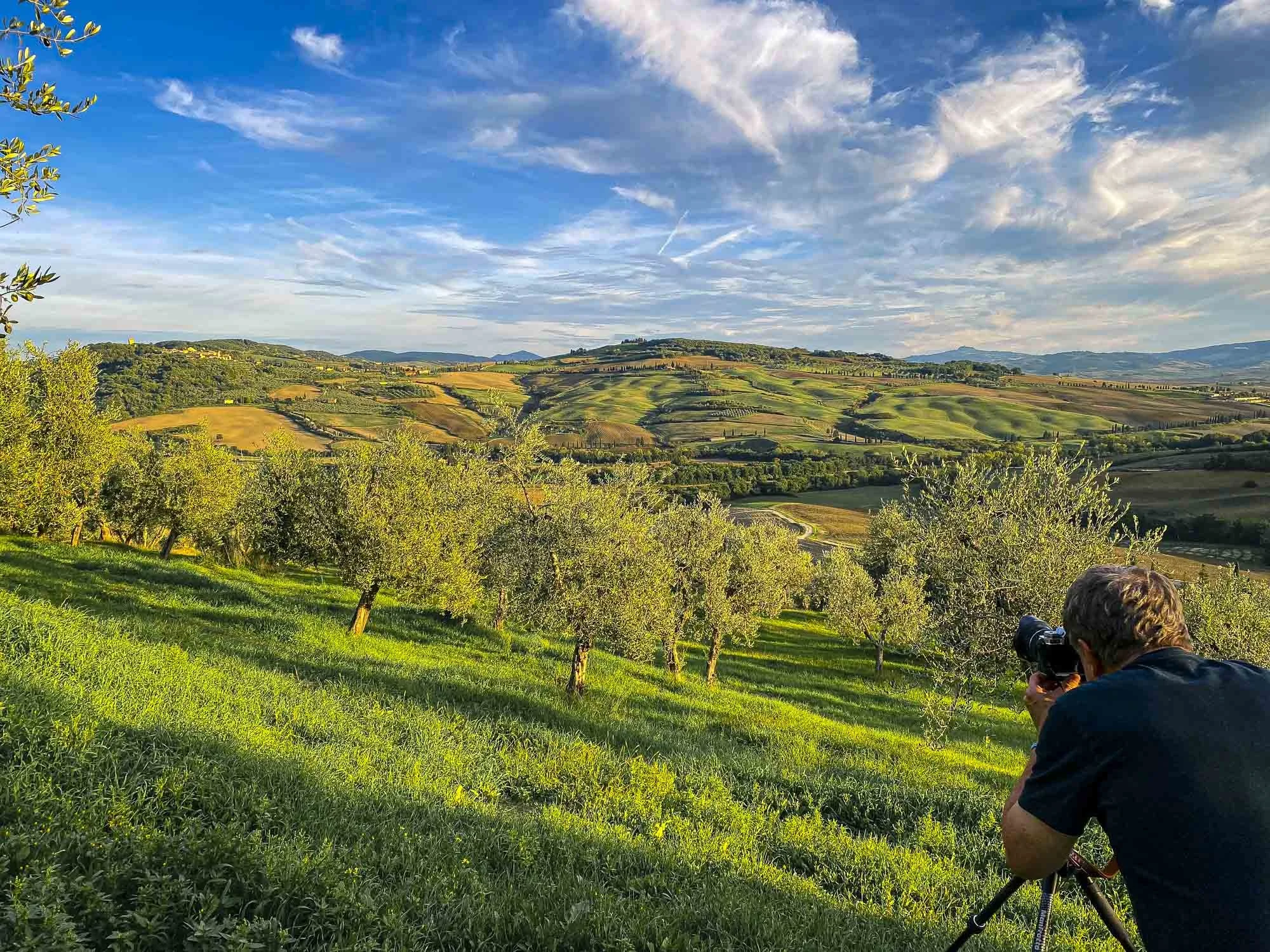 A person with brown hair taking a photograph of a landscape with a camera on a tripod, overlooking a lush green valley with trees and rolling hills under a partly cloudy sky.