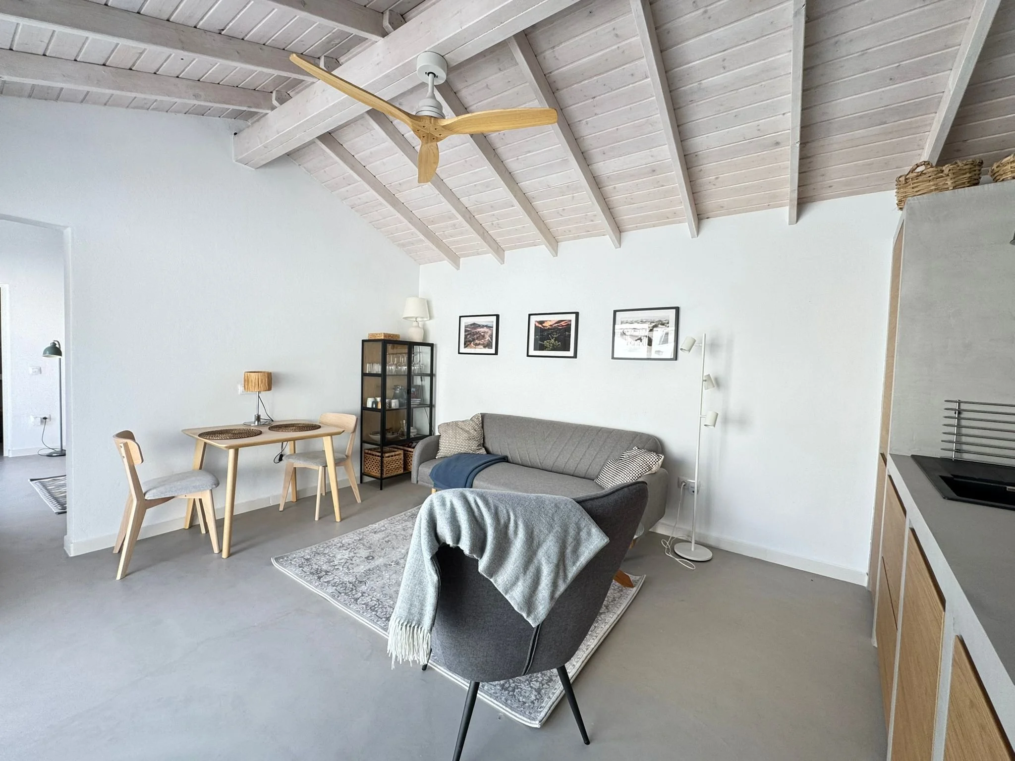 Interior of a living room with a vaulted ceiling, light wood beams, and a ceiling fan. The room features a gray sofa, a black glass display cabinet, a round dining table with four chairs, and decorated with framed pictures on the wall. There is a whi