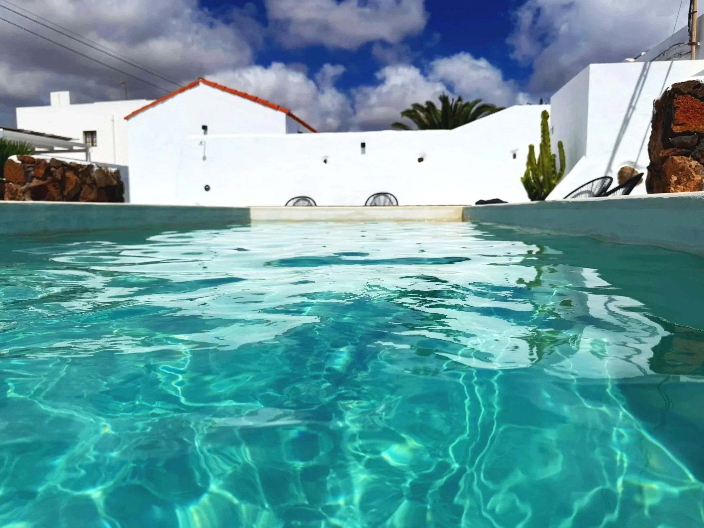 View of a swimming pool with turquoise water in the foreground, surrounded by white buildings topped with red tiles, against a backdrop of a partly cloudy blue sky.