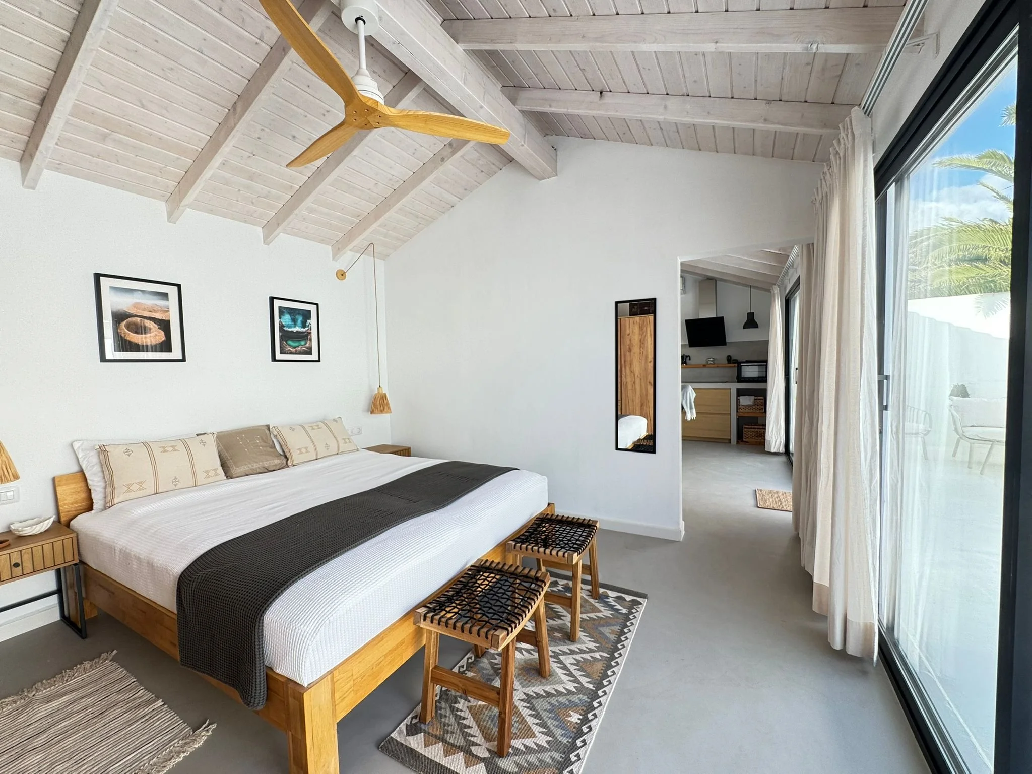 A bedroom with a wooden bed frame, white bedding, and brown and beige pillows. There are two black-framed pictures on the white wall above the bed. Three small wooden stools are placed at the foot of the bed on a patterned rug. A pendant light hangs 
