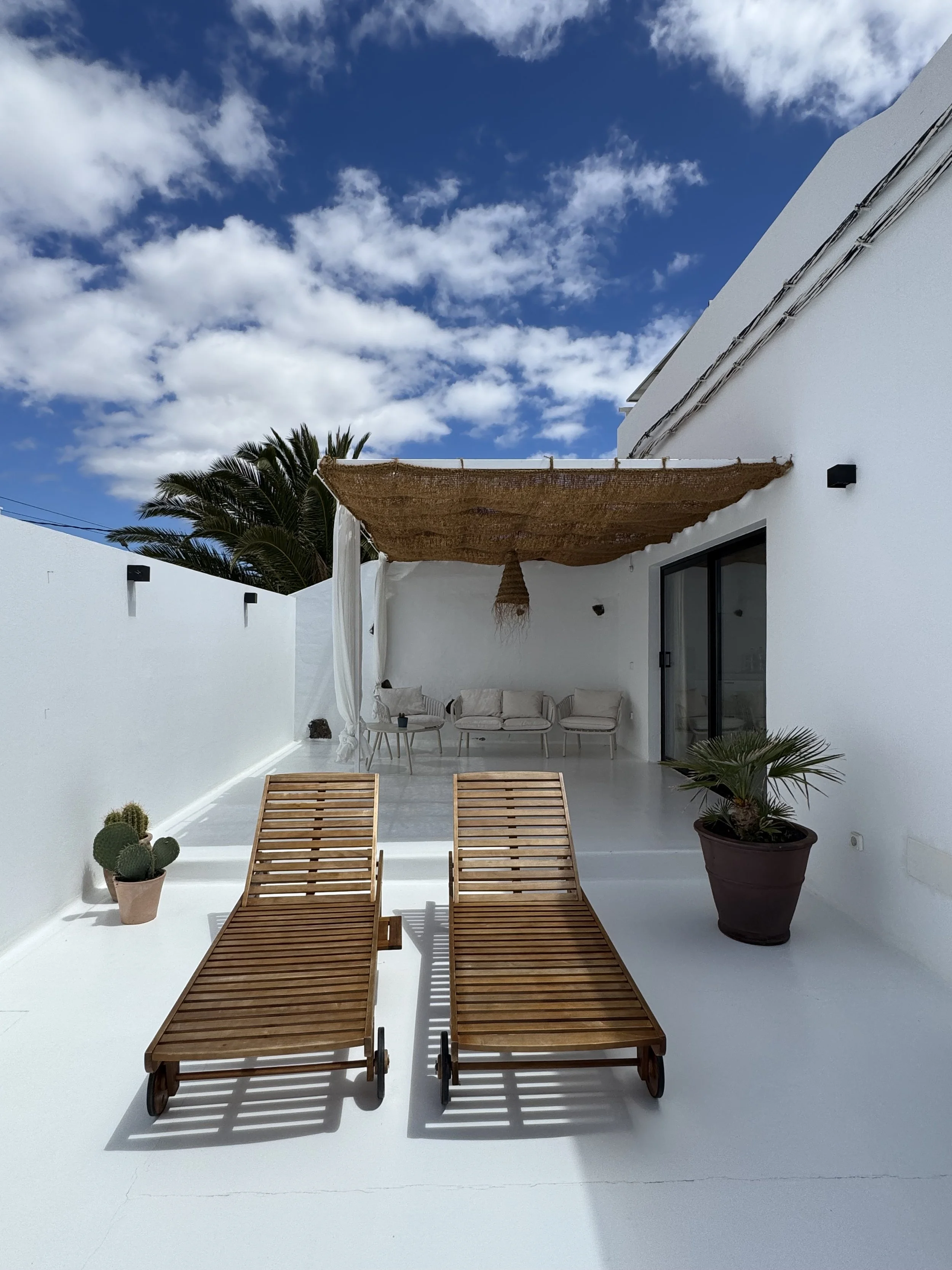 A white outdoor patio area with two wooden lounge chairs, potted plants, a white sectional sofa under a shade, and a partly cloudy blue sky.