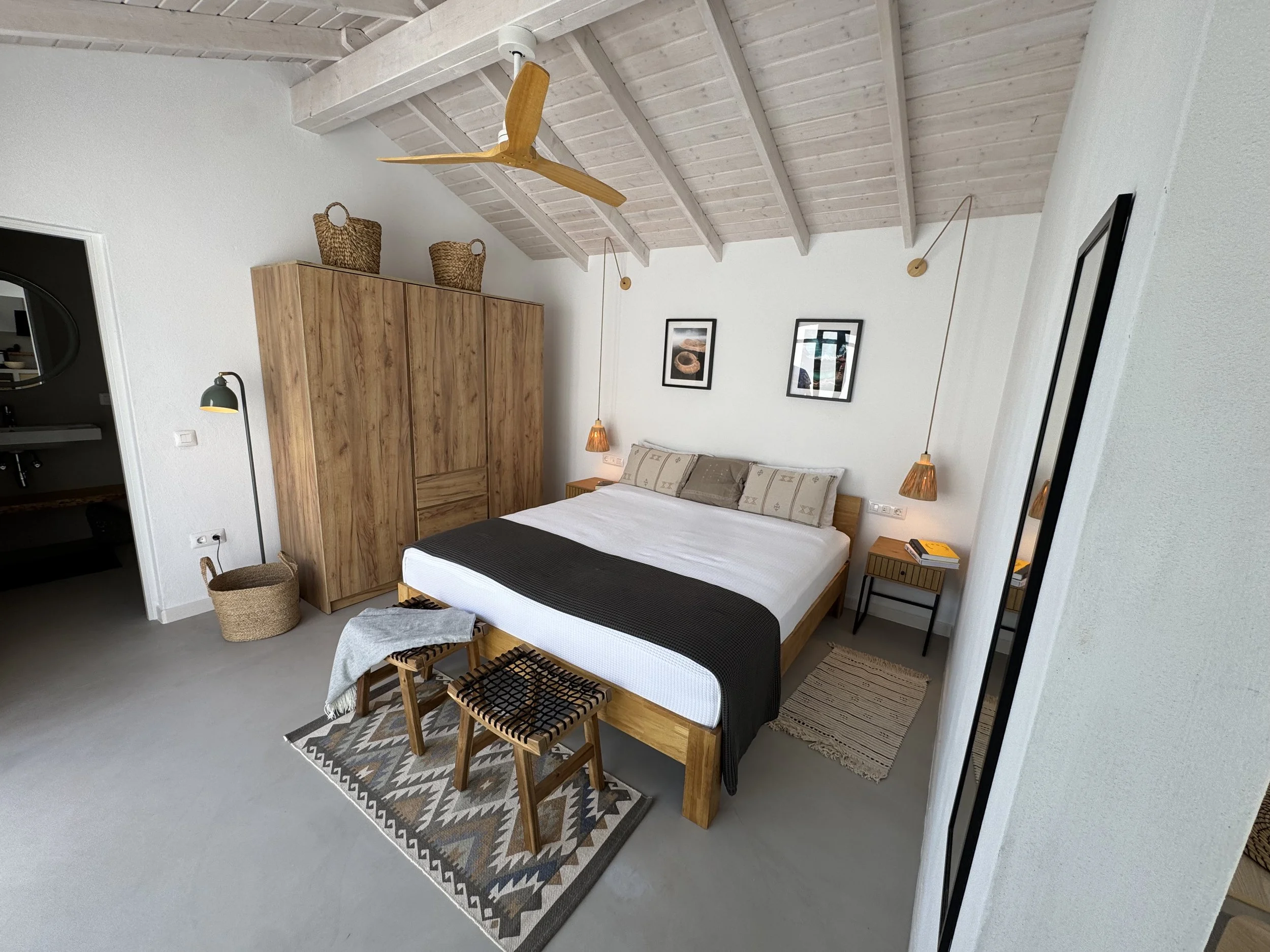 A bedroom with a wooden bed, patterned rug, and wooden accents. There are two stools at the foot of the bed, a wardrobe with woven baskets on top, and framed artwork on the walls. The ceiling is made of light wood with exposed beams and a ceiling fan