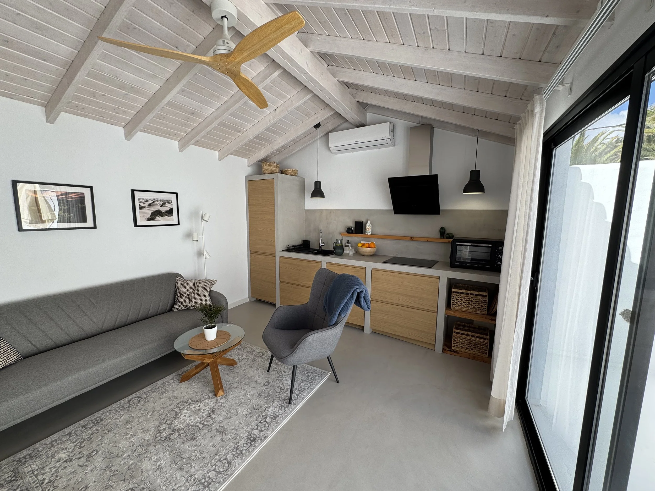 Interior view of a modern living room with a kitchen area, featuring a gray sofa, a gray armchair, a wooden coffee table, a white wall with two framed photos, a ceiling fan, air conditioner, pendant lights, and large sliding glass doors with curtains