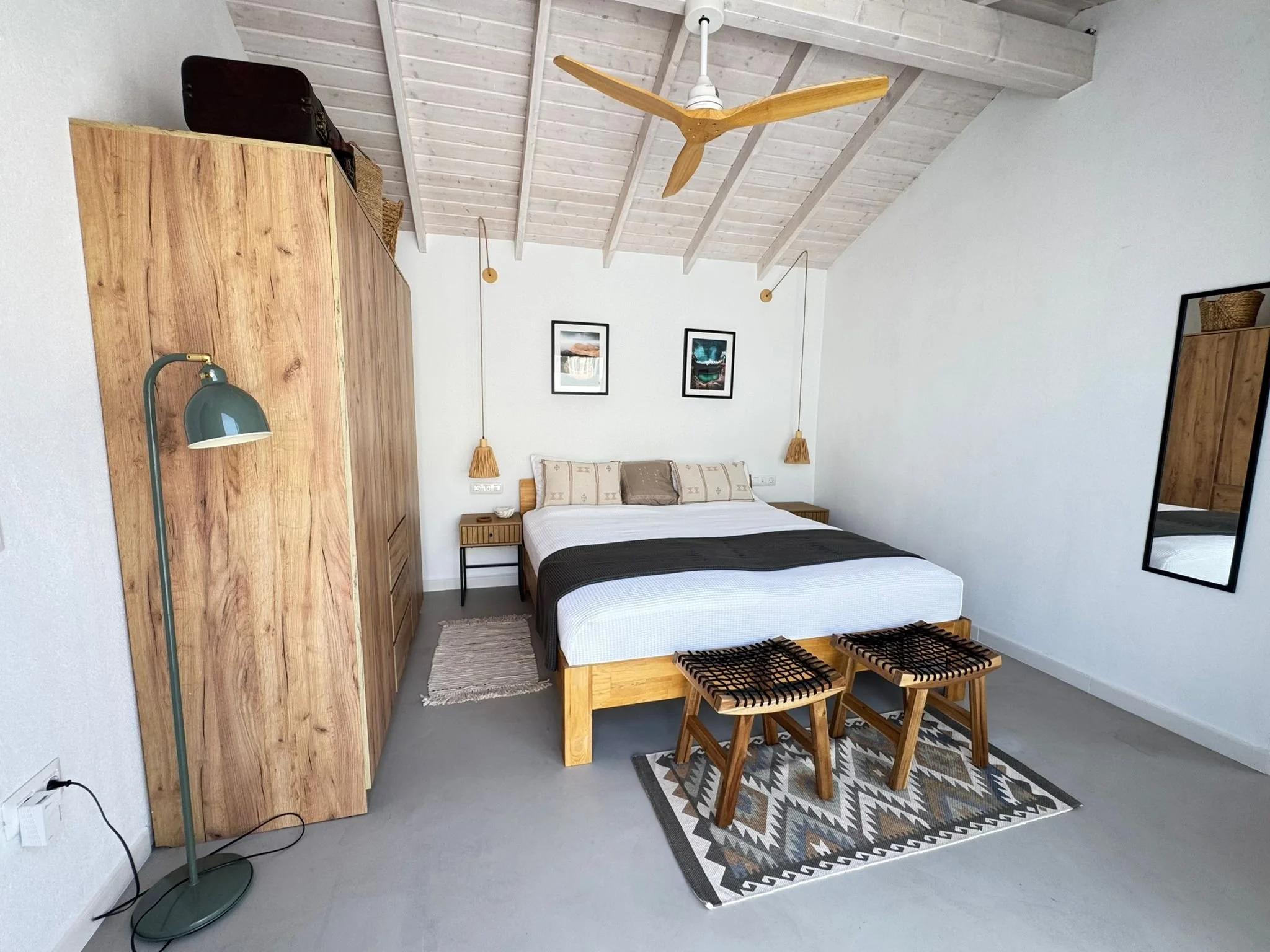 A bedroom with a wooden wardrobe, a bed with pillows, a side table, a mirror, a colorful patterned rug, and a wooden bench. The ceiling has white-painted wooden beams and a ceiling fan with wooden blades. Two hanging pendant lights with straw shades 