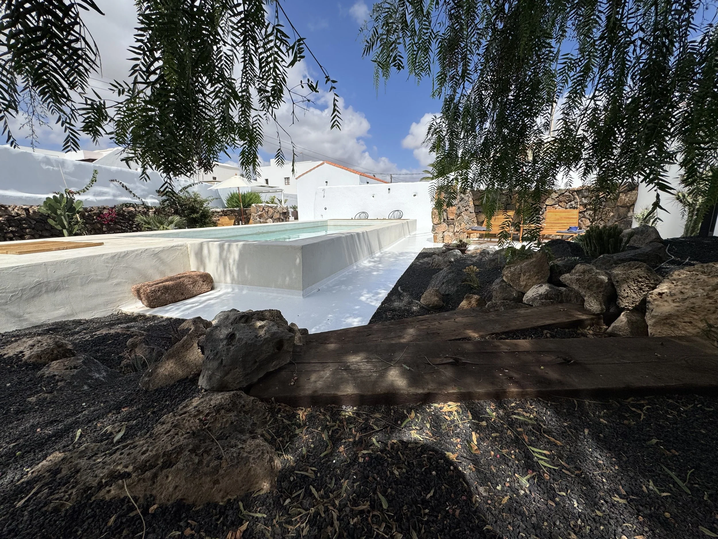 A courtyard with a small pool, surrounded by white walls, with wooden benches and plants. The photo is taken from a shaded area with rocks and a wooden plank in the foreground, looking towards the open sky with clouds.