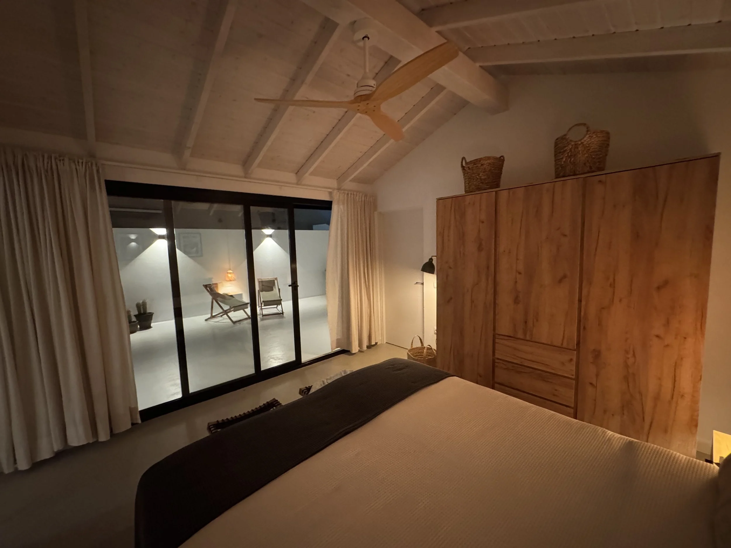 A cozy bedroom with a wooden ceiling fan, large window with beige curtains, and a sliding glass door leading to an outdoor sitting area with chairs and a small lamp.