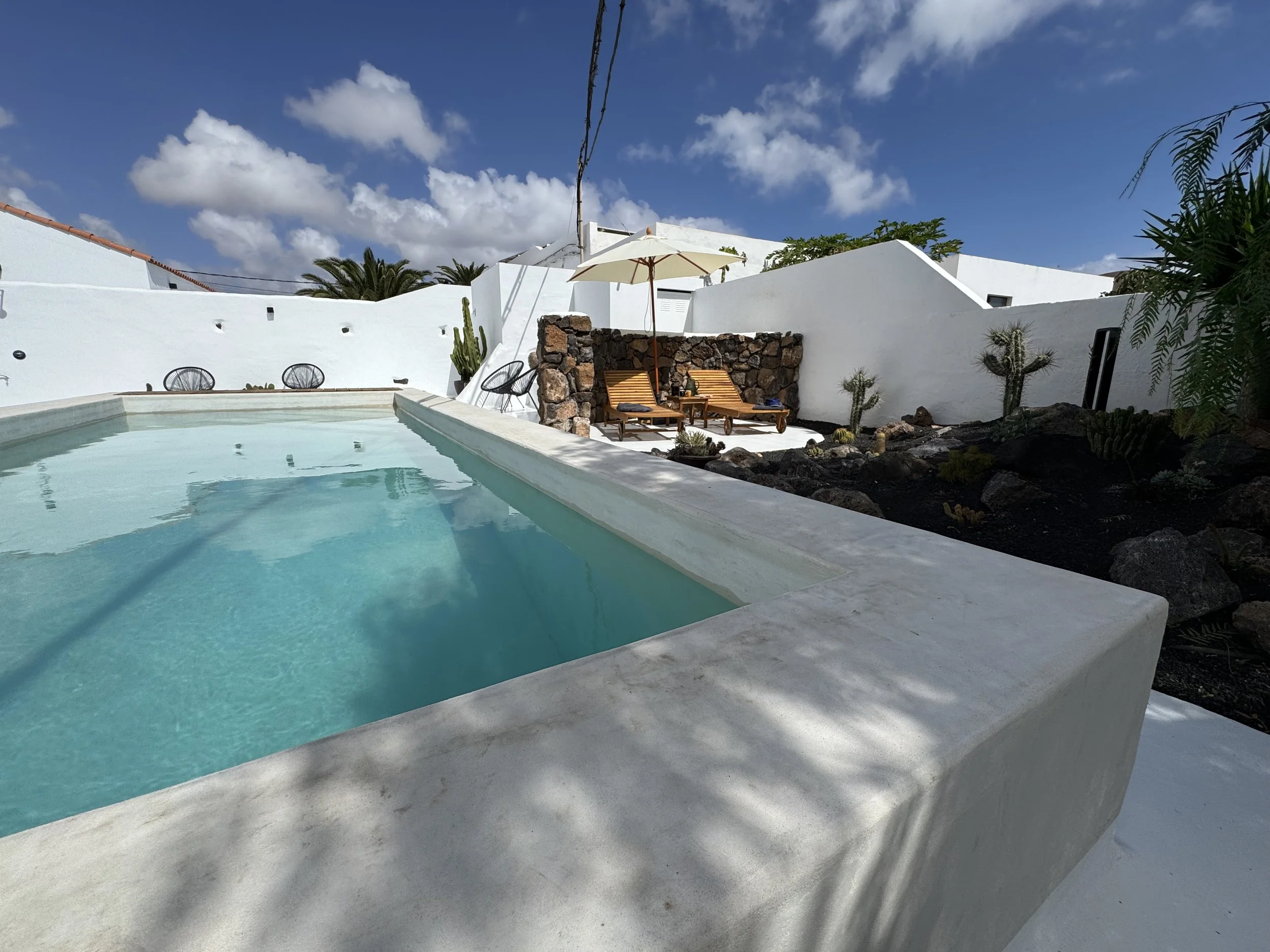 A swimming pool with turquoise water, surrounded by whitewashed walls, desert plants, and outdoor seating with an umbrella.