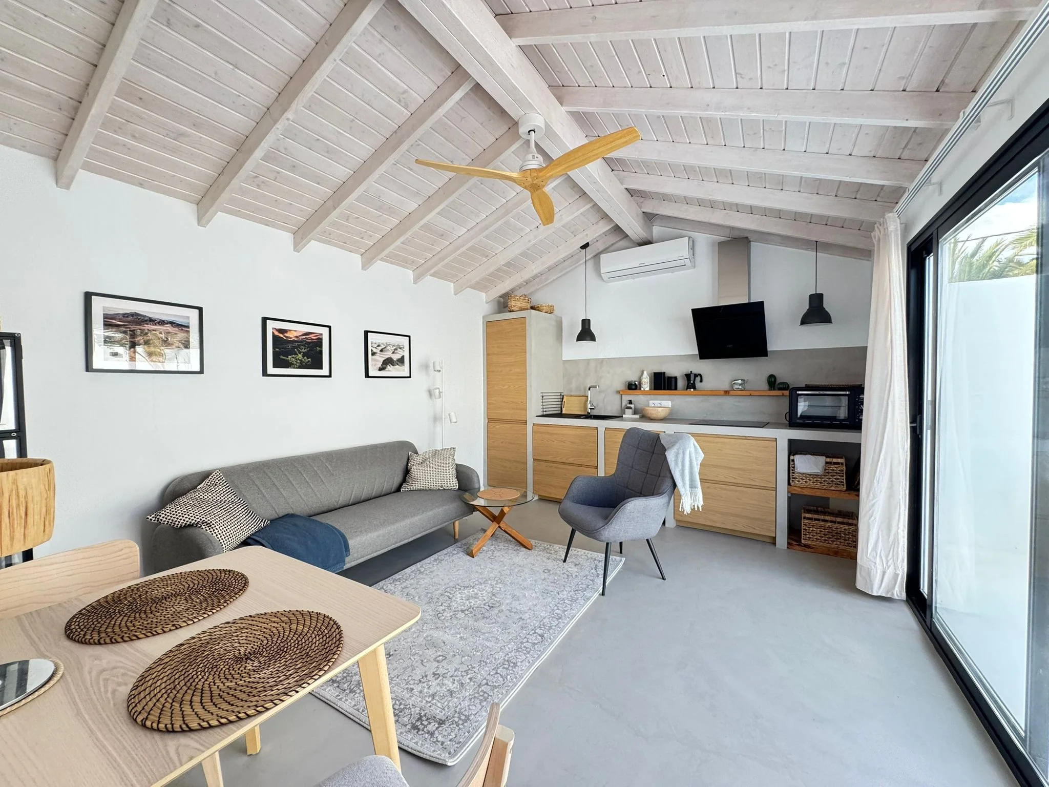 A modern living room with a vaulted wooden ceiling, a ceiling fan, a gray sofa with patterned pillows, a dining table with woven placemats, a small gray armchair, framed pictures on the wall, and a kitchen area with cabinets, a microwave, and a TV, w