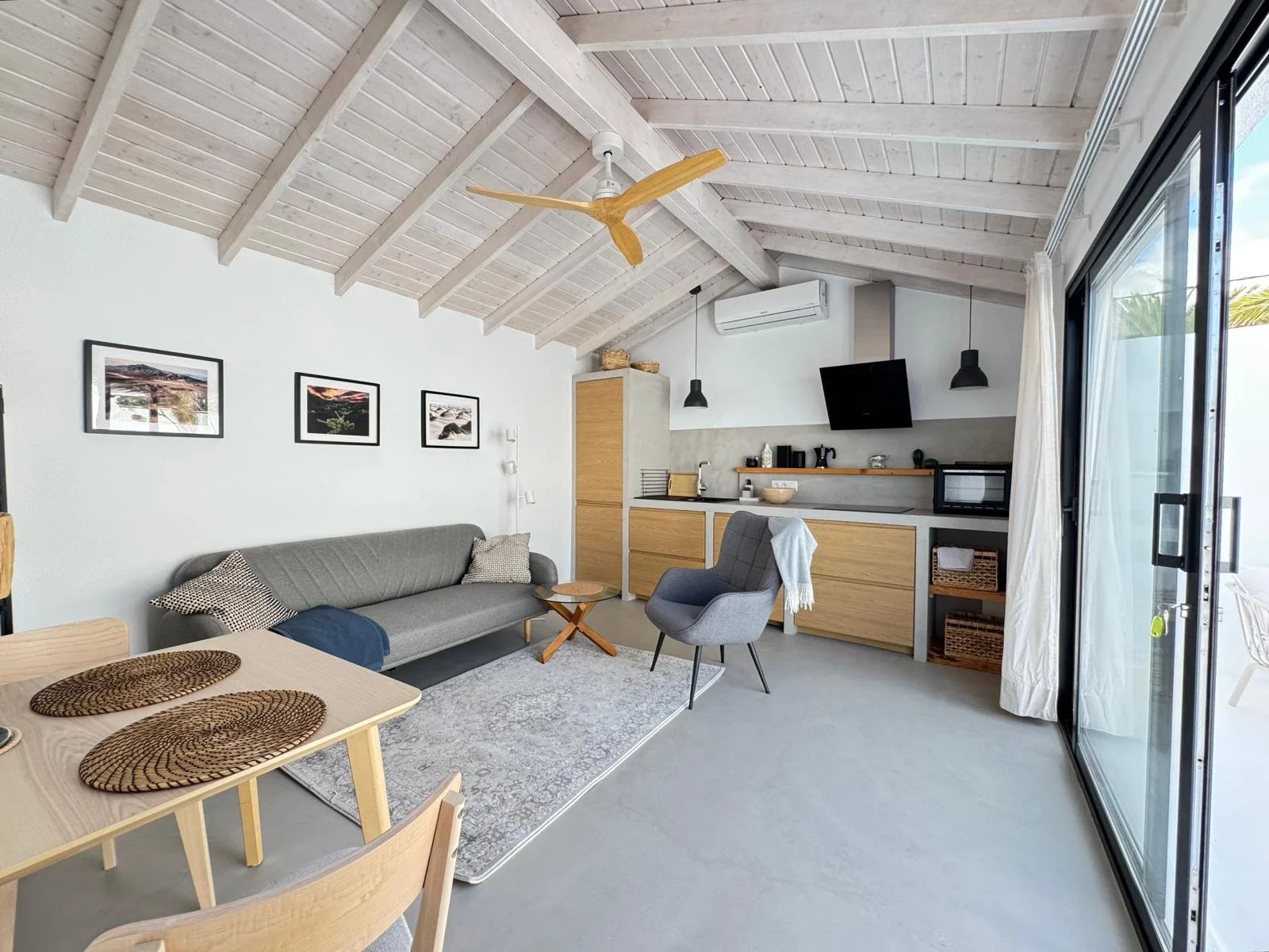 Living room and kitchen with vaulted white wooden ceiling, gray sofa, gray armchair with white throw, dining table with woven placemats, sliding glass door with white curtains, wall art, black TV, compact kitchen with wooden cabinets and open shelving, and outdoor patio visible through sliding door.