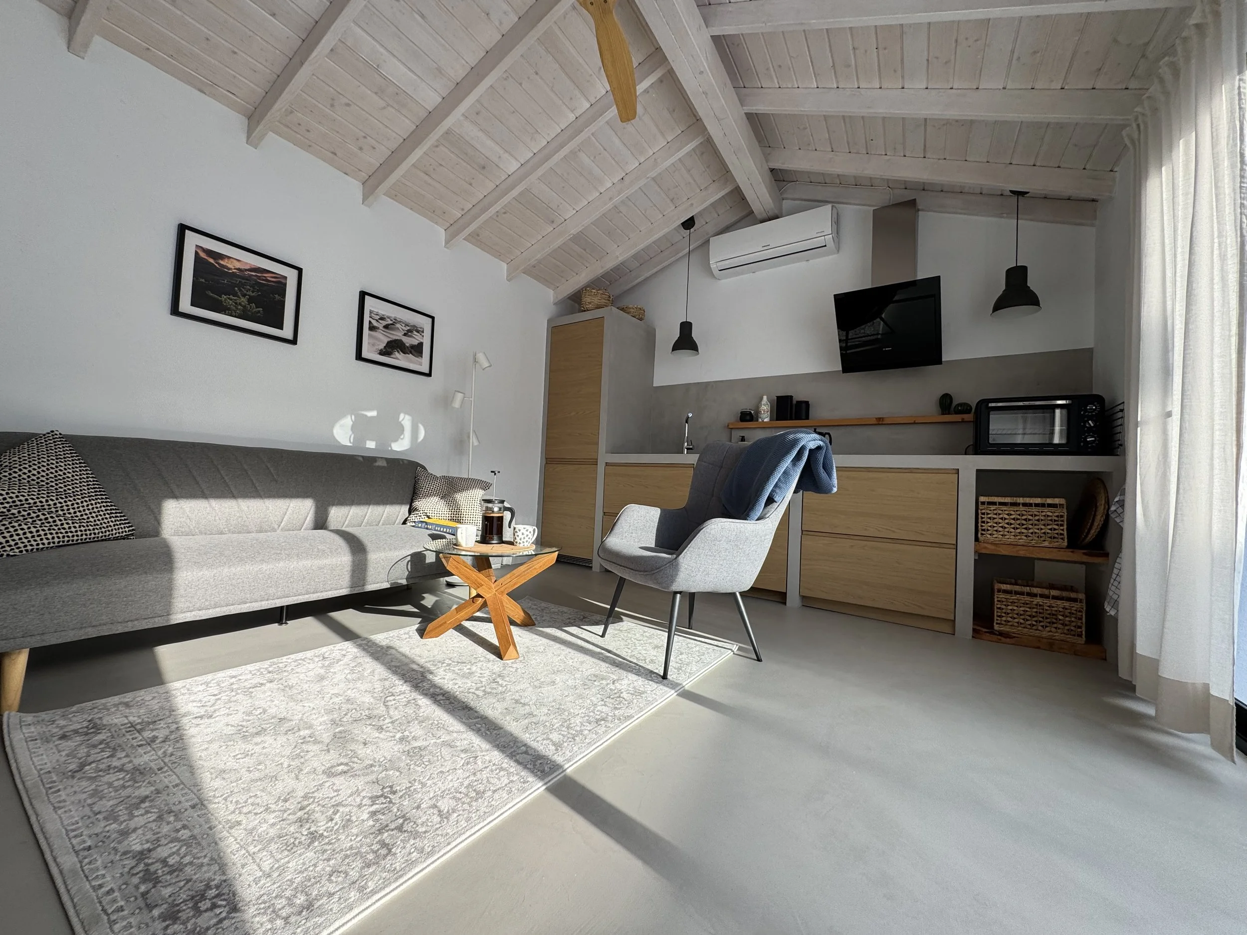 Living room with a gray sofa, a small wooden coffee table with drinks, two framed landscape pictures on the wall, a gray and white area rug, a light gray armchair with a blue throw, a white wall-mounted air conditioner, black pendant lights, and a ki