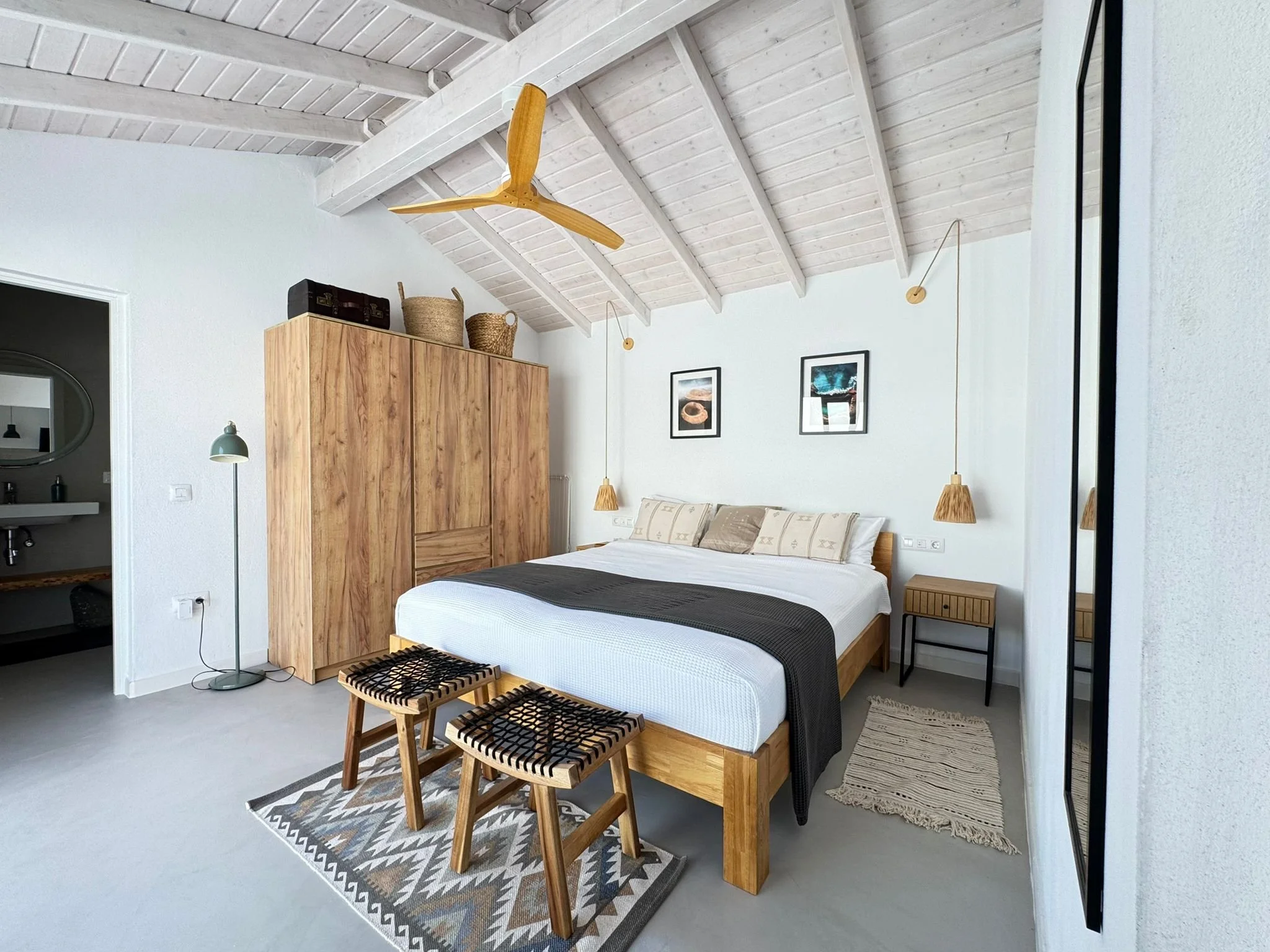 A bedroom with white walls, a vaulted ceiling with exposed beams painted white, a wooden wardrobe, framed artwork, a bed with a beige headboard, and woven footstools. There is a standing lamp and hanging pendant lights with woven shades.