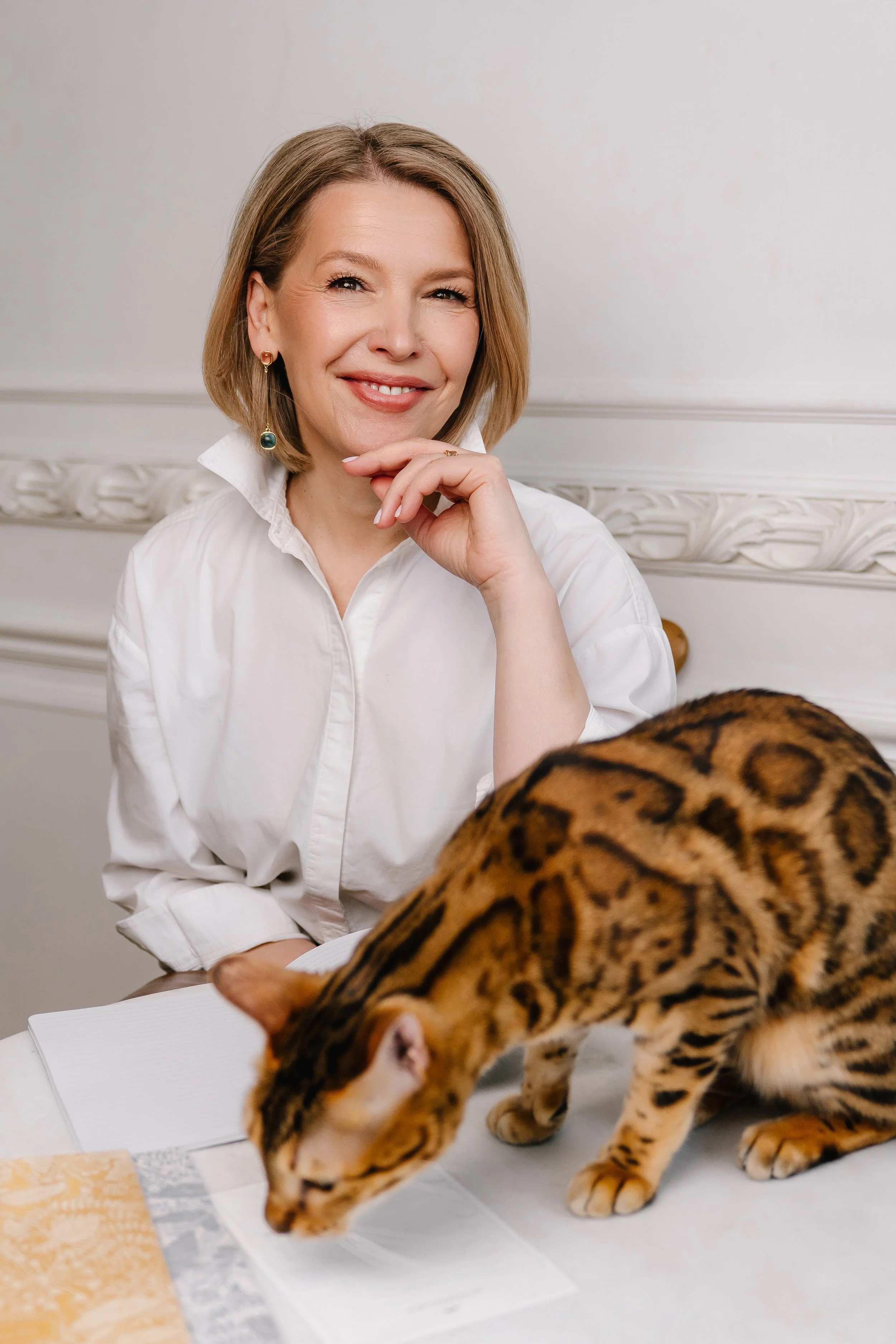 A woman with blonde hair in a white shirt, smiling thoughtfully, sitting at a table with a notebook and maps. She is touching her chin with her right hand, and a Bengal cat is walking across the table in front of her.