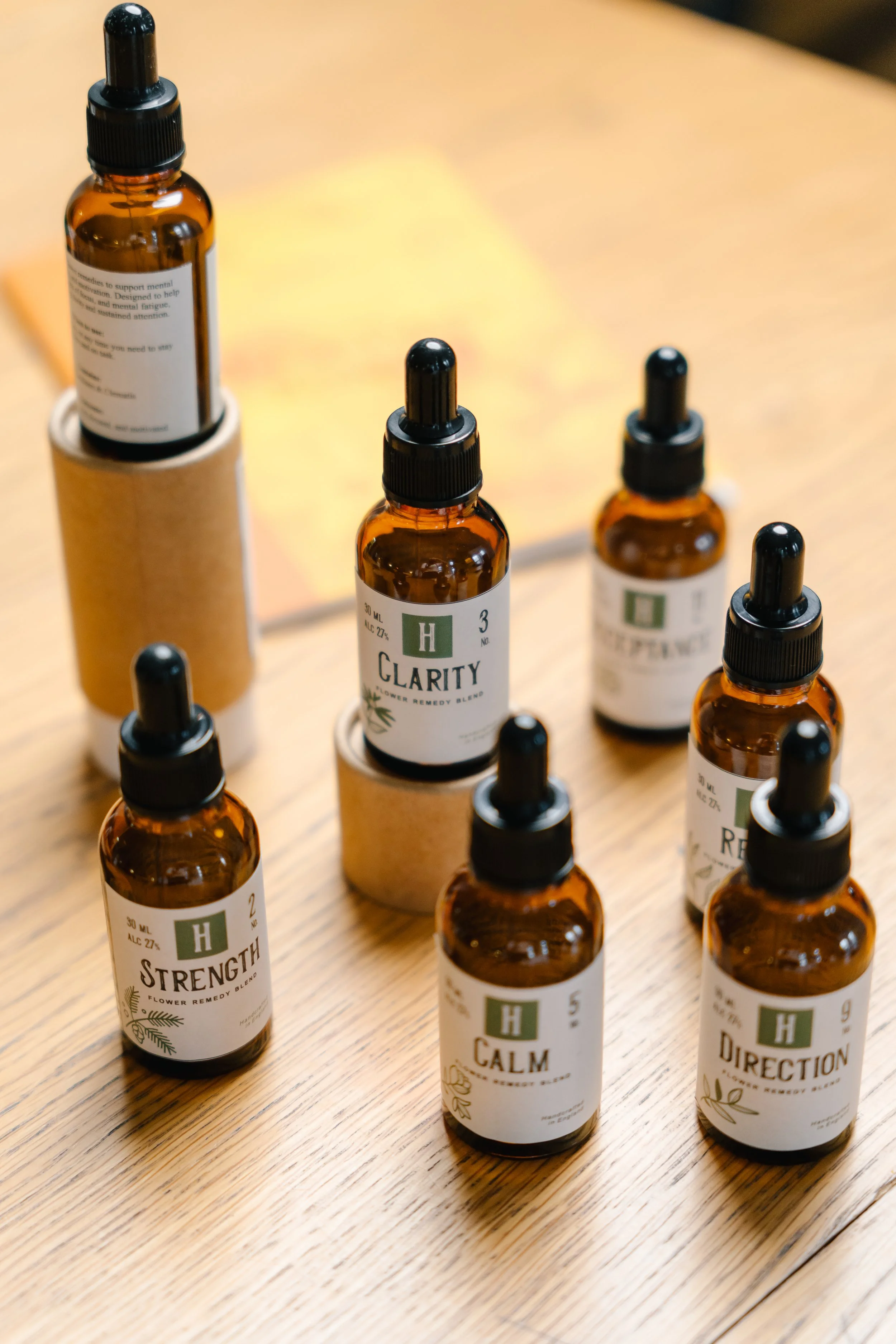 Several small amber glass bottles with black dropper caps are arranged on a wooden surface. The bottles have white labels with green and black text indicating they contain herbal remedy blends for various purposes like strength, calm, and direction.