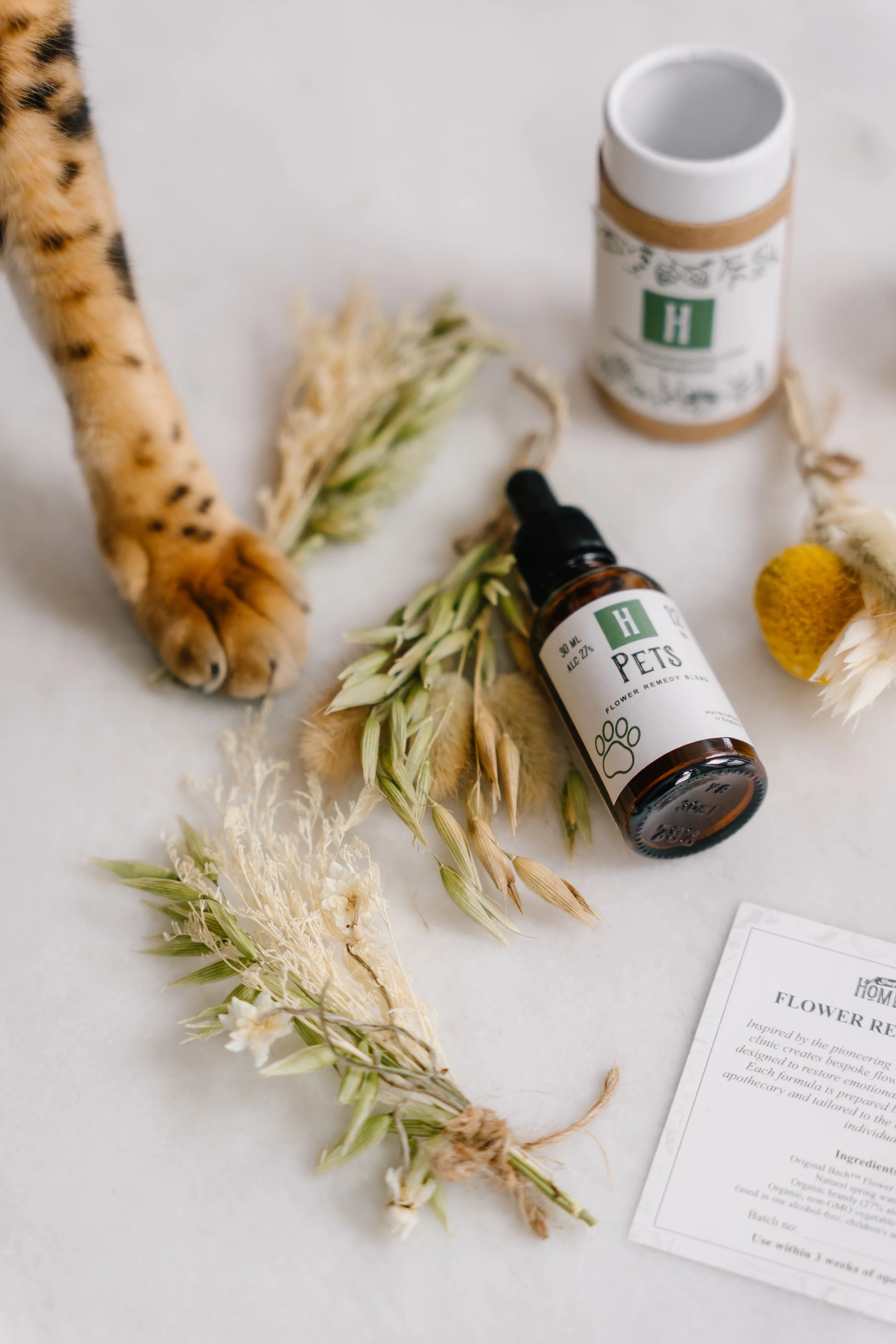 Dried herbs and flowers, a small brown bottle labeled 'Pets Flower Remedy Blend' with a dropper cap, a white ceramic container with a green label, and some papers on a white surface.