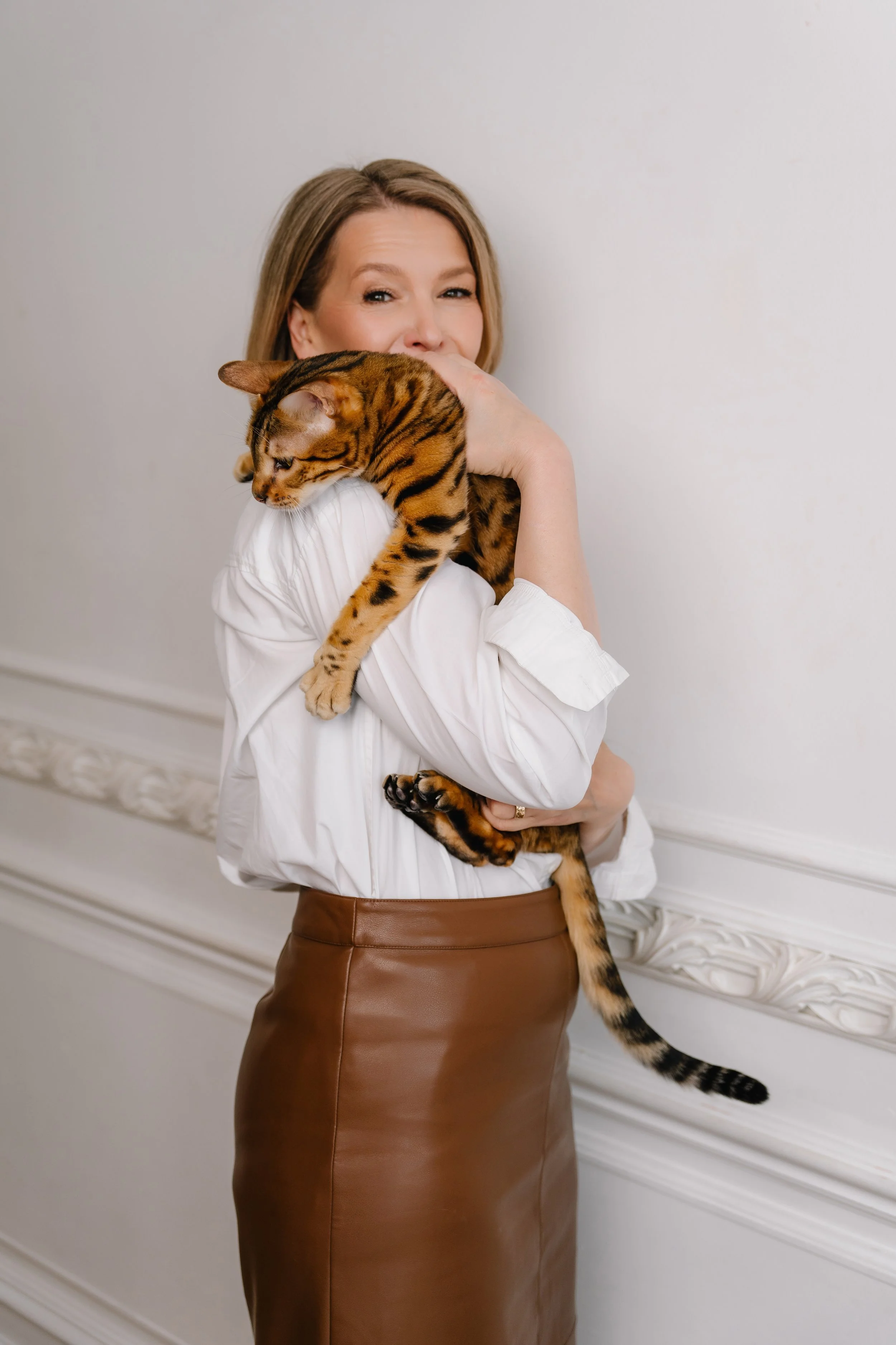 A woman with blonde hair wearing a white shirt and brown leather skirt, smiling while holding a Bengal cat with a spotted coat and long tail, standing against a plain light-colored wall.