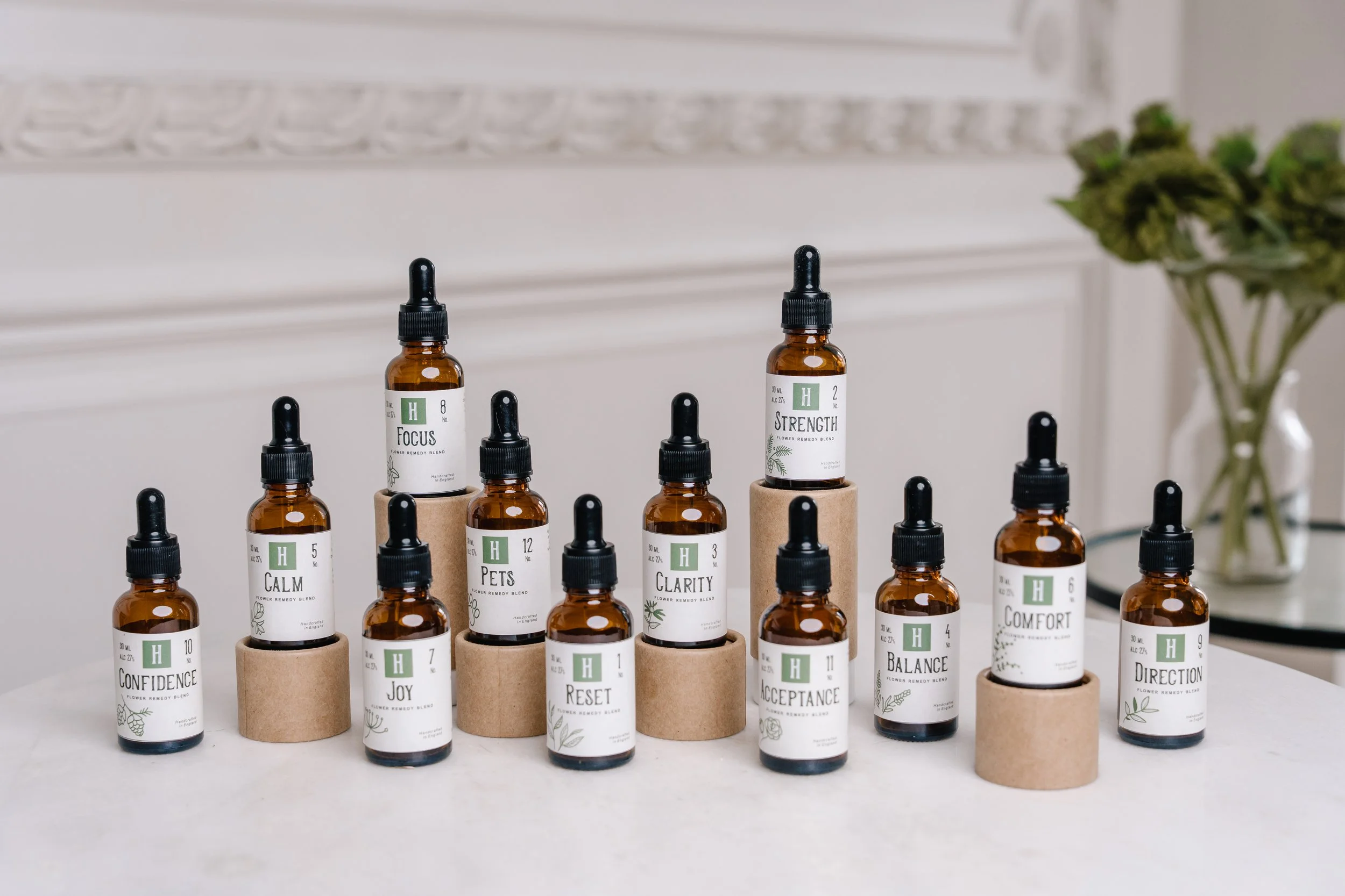 A display of small amber glass bottles with black dropper caps, labeled with various names related to mental states and feelings, such as "Focus," "Calm," "Confidence," and "Joy." The bottles are positioned on circular cardboard stands, arranged in rows on a white surface, with a blurred background that includes a white wall with decorative molding and a vase of green flowers.