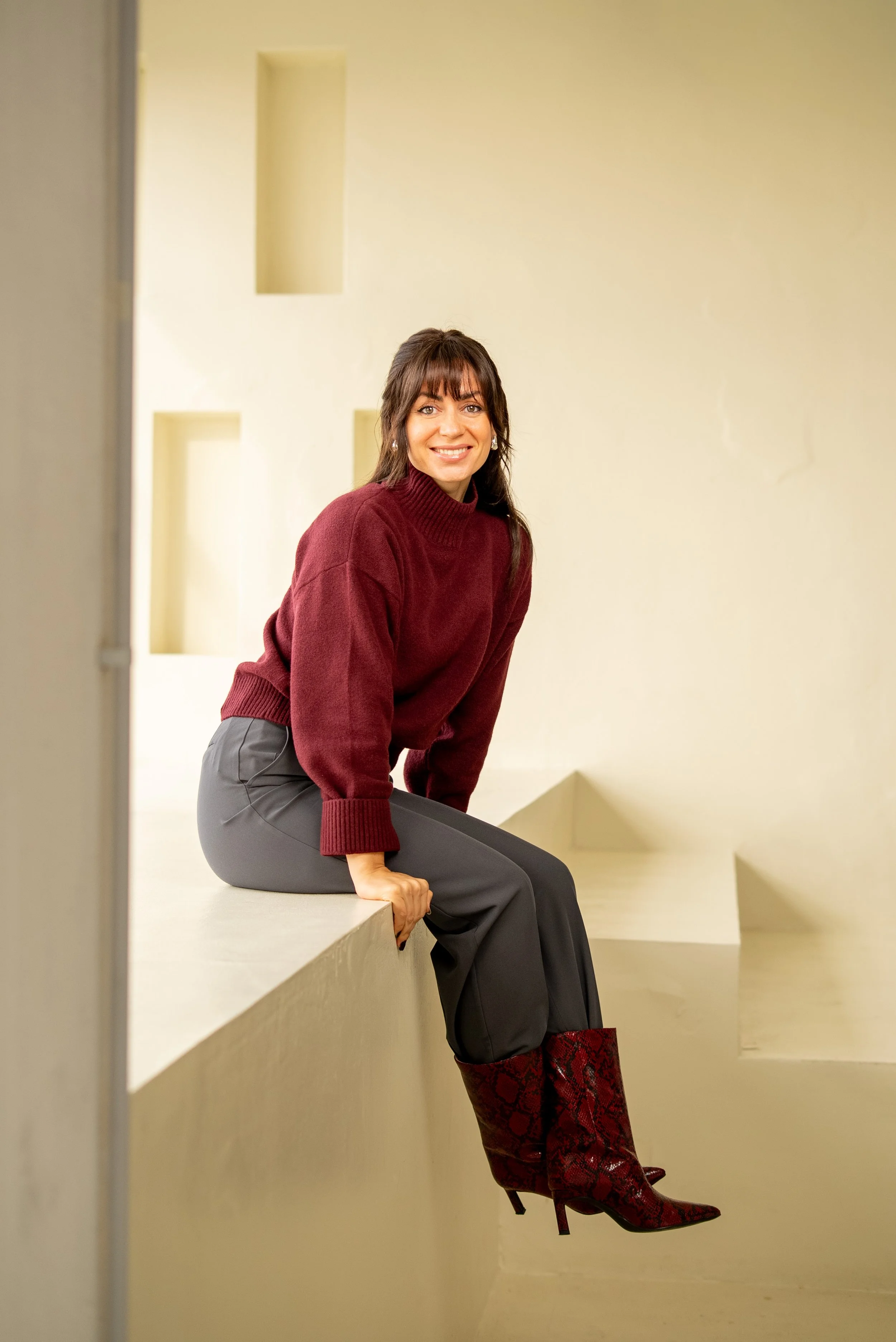A woman sitting on a ledge, wearing a burgundy sweater, gray pants, and red snake-print high-heeled boots, smiling at the camera inside a modern, minimalist interior.