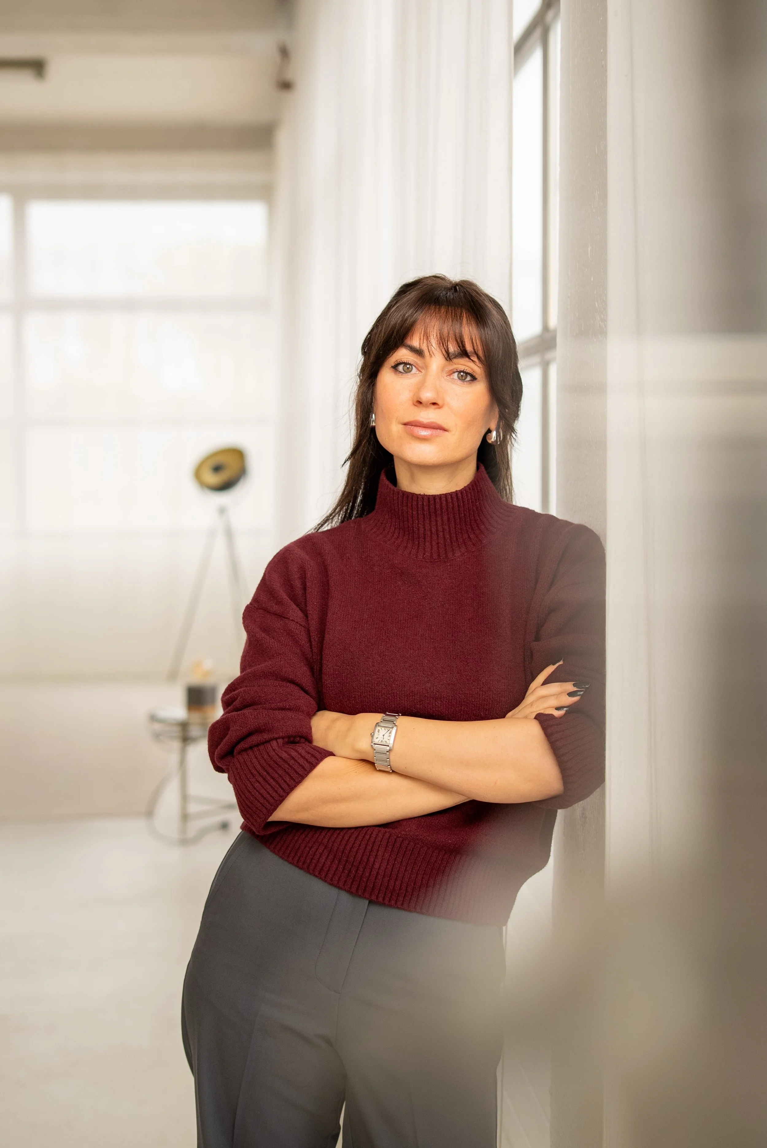 A woman with dark brown hair and bangs, wearing a maroon turtleneck sweater, stands with arms crossed against a white wall in a bright room with large windows and a lamp in the background.
