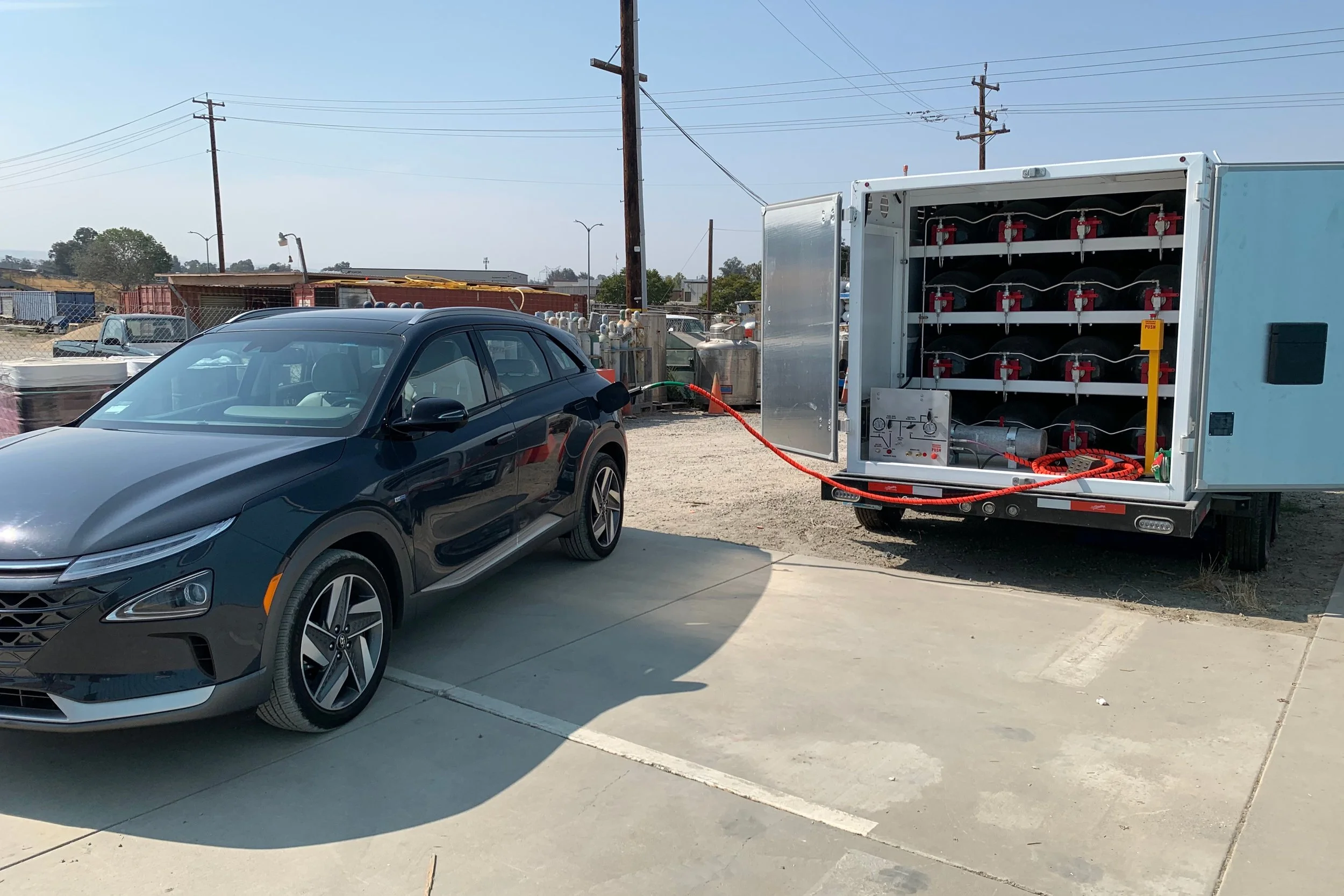 A black car connected to a mobile hydrogen transport trailer by a red cable on a concrete parking lot.