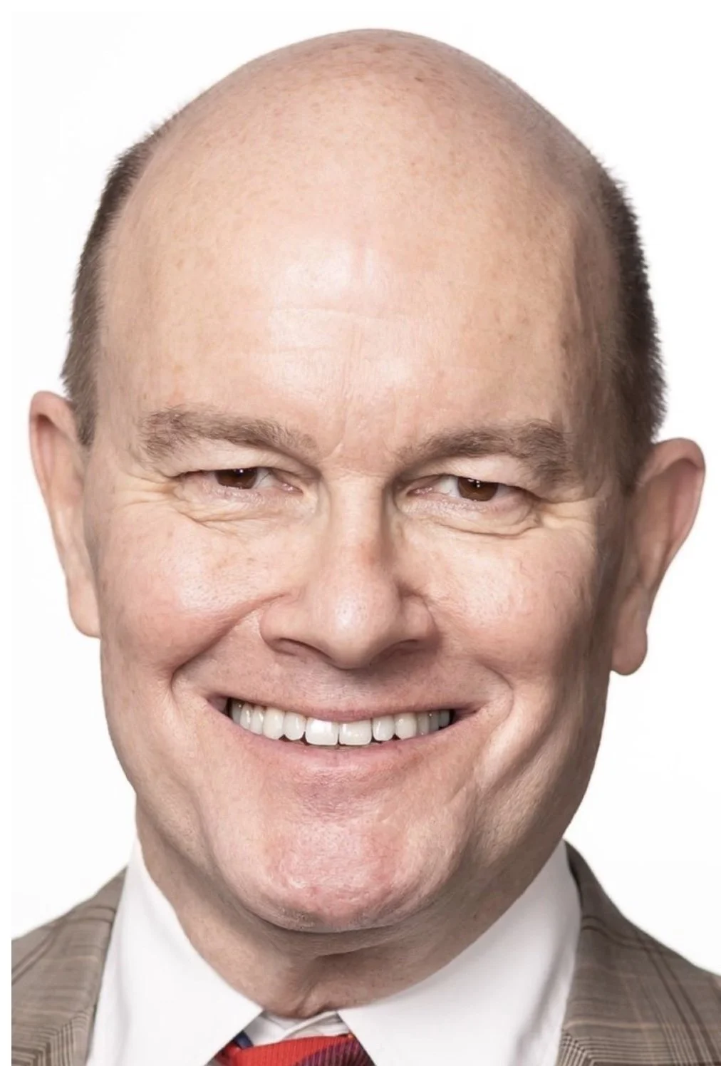 A close-up portrait of a man with a shaved or bald head, light skin, and a wide smile, wearing a plaid suit jacket, white shirt, and red tie.
