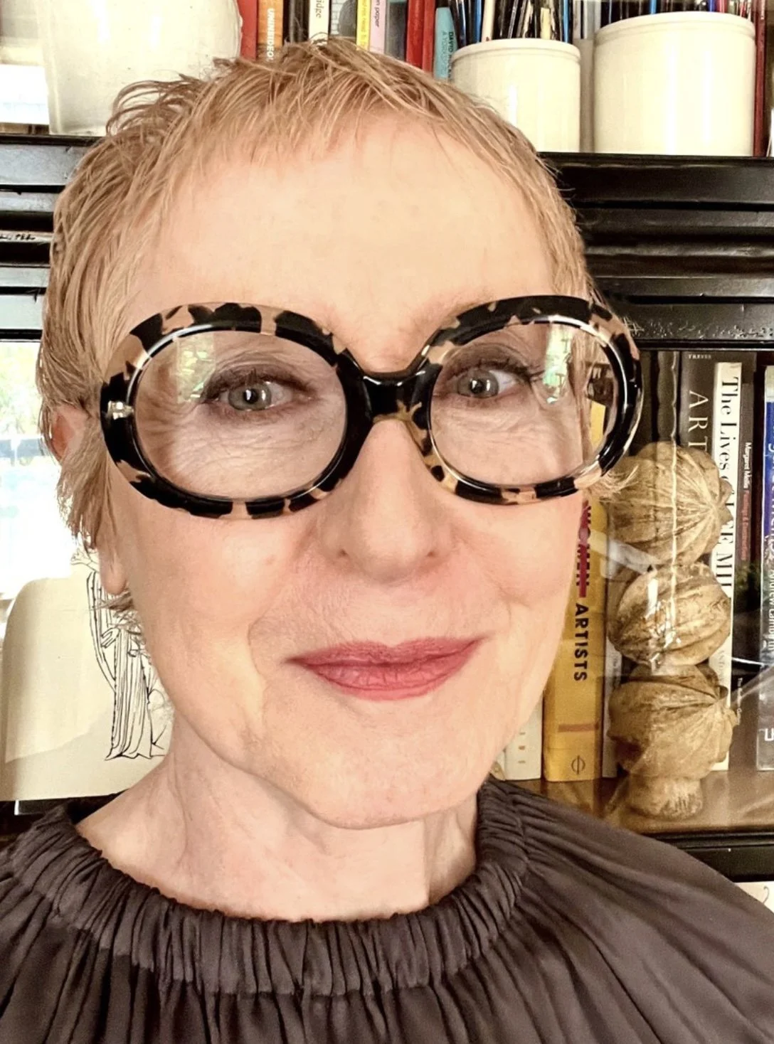 A woman with short blonde hair is wearing large tortoiseshell glasses and has a slight smile. Behind her, there are shelves with books, a small illustration, and a bundle of decorative objects.