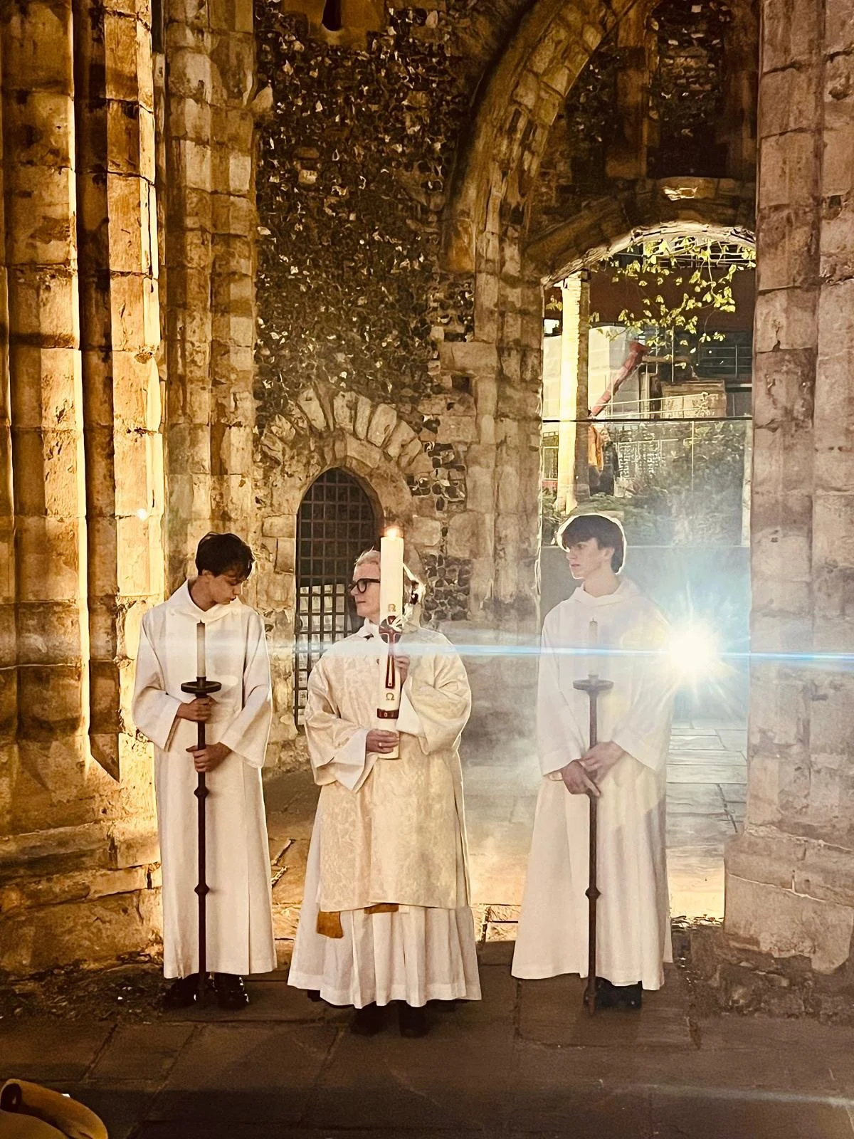 Three young men dressed in white robes, holding candles, standing inside a stone church with arched doorways and ancient stone walls.