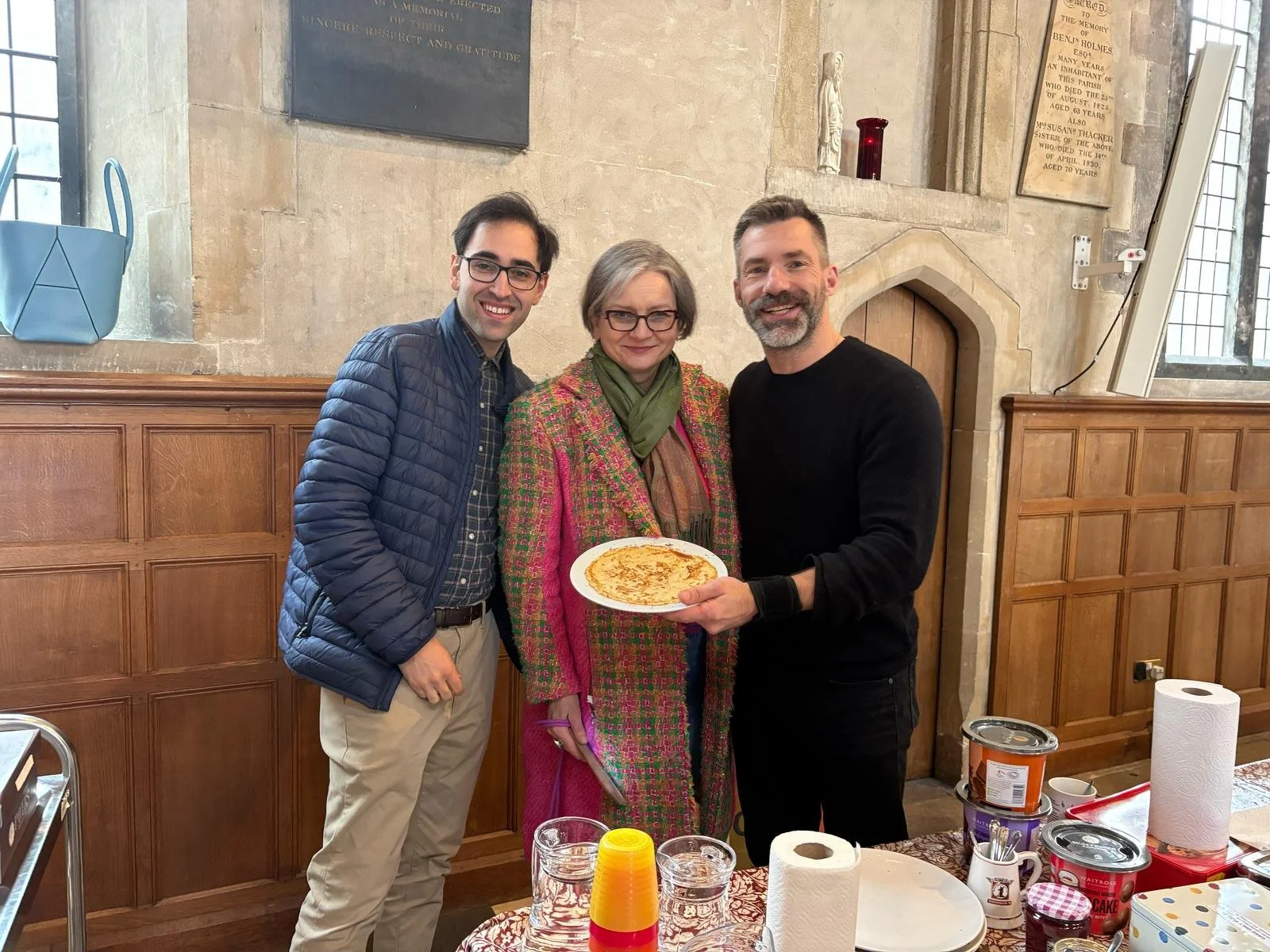 Three people standing close together in a room with wooden paneling and stone walls, smiling at the camera. The person in the middle is holding a plate with a pancake. The table in front has paper towels, cups, and food containers, suggesting a gathe