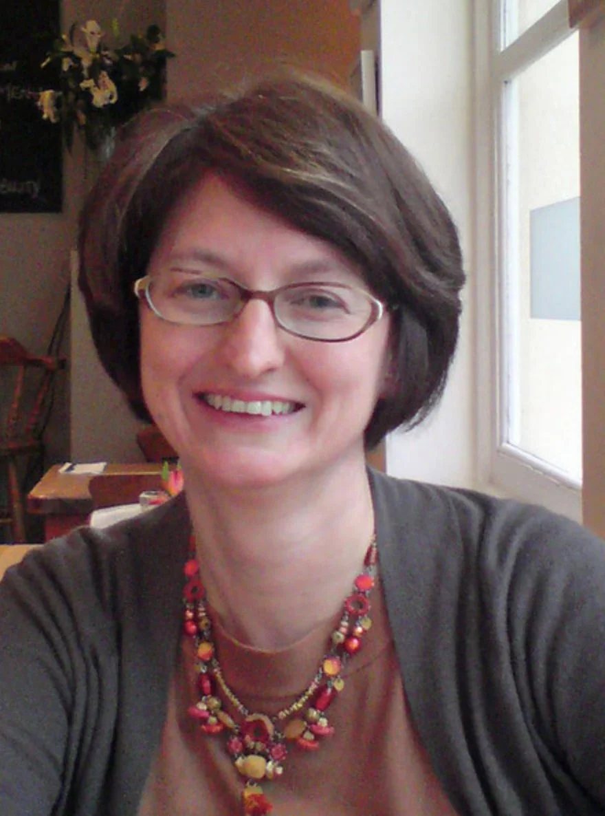 A smiling woman with short dark hair wearing glasses, a gray cardigan, and a red, yellow, and orange beaded necklace, sitting near a window with a wooden table and floral decoration in the background.