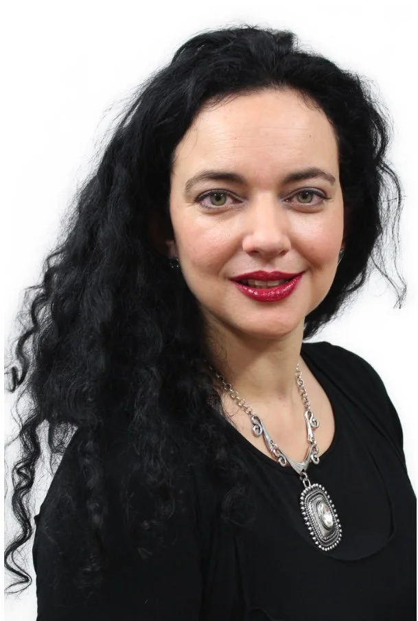 A woman with long, curly black hair, fair skin, wearing a black top and a silver necklace, smiling, against a plain white background.