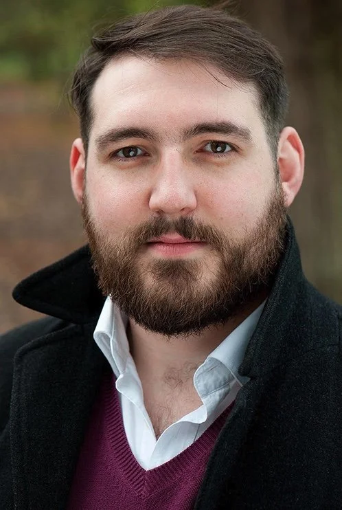 A close-up portrait of a young man with a beard, wearing a white shirt, purple sweater, and black coat outdoors.