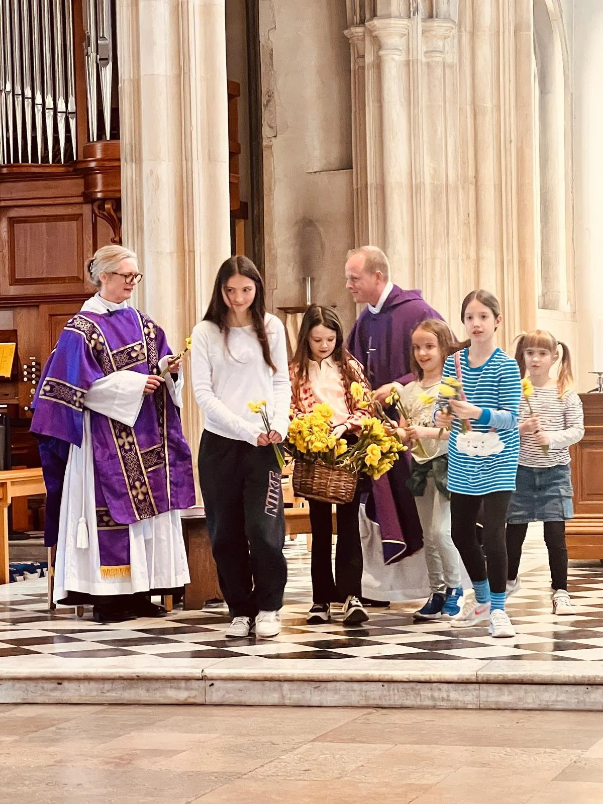 Children and two clergymen in church holding yellow flowers, inside a church with large columns and checkered floor.