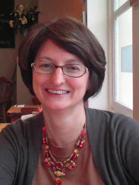 Smiling woman with short brown hair, glasses, wearing a multicolored beaded necklace and gray sweater, sitting at a table near a window in a cozy room.