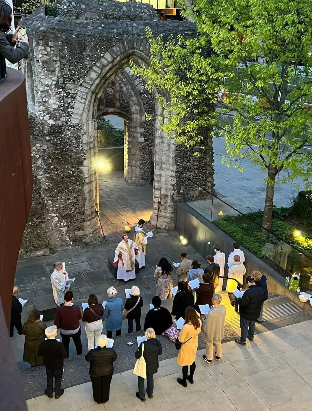 Group of people gathered outdoors at an event near ancient stone ruins with a large arched window. Some individuals are wearing robes, possibly clergy, and others hold papers. There are trees and modern glass railings in the background, and the area