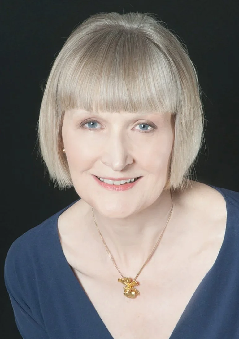 A woman with short blond hair and blue eyes smiling, wearing a blue top and a gold necklace with a rabbit pendant, against a black background.