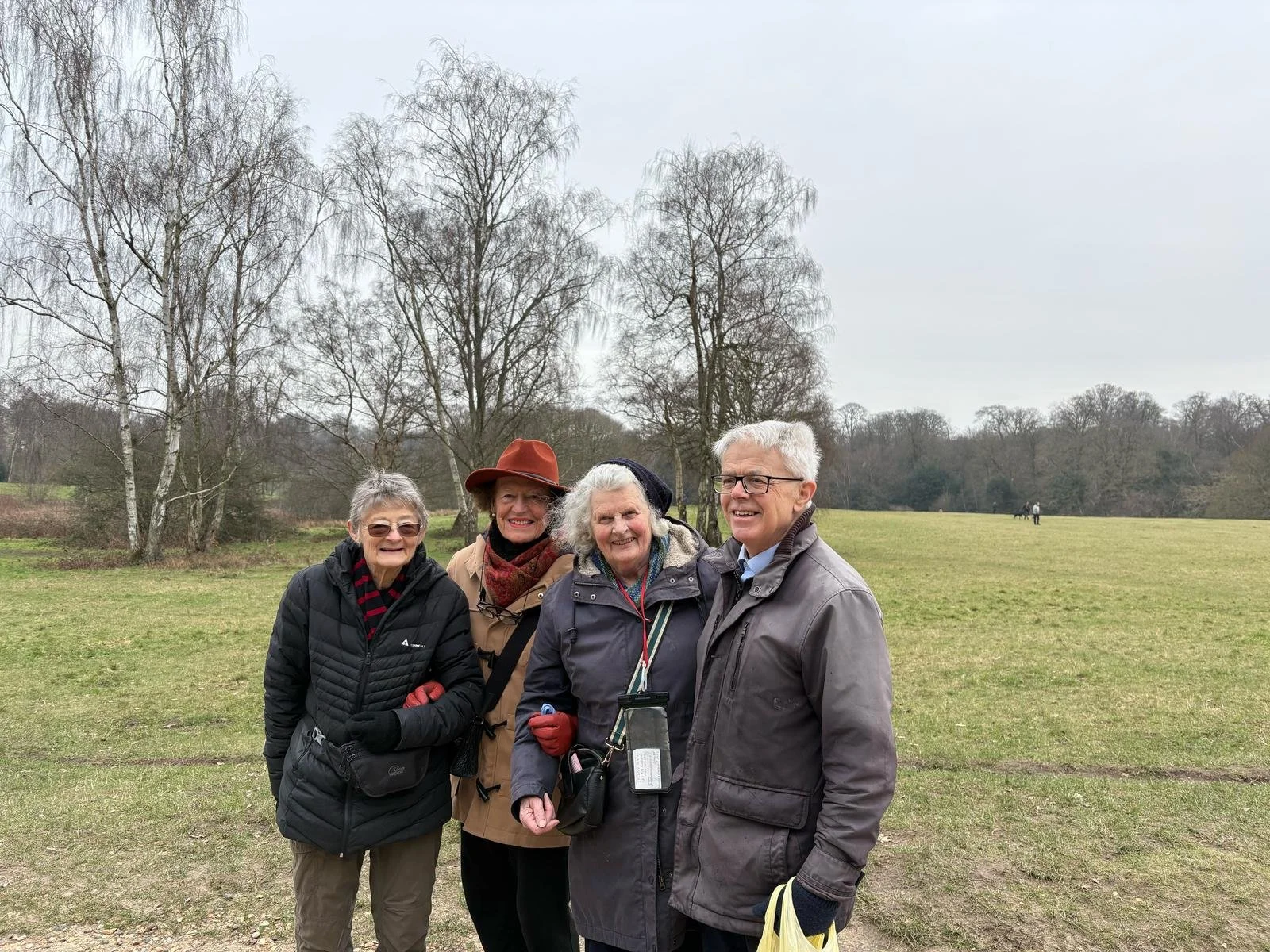 Four elderly people smiling and standing together outdoors in a field with bare trees in the background on an overcast day.
